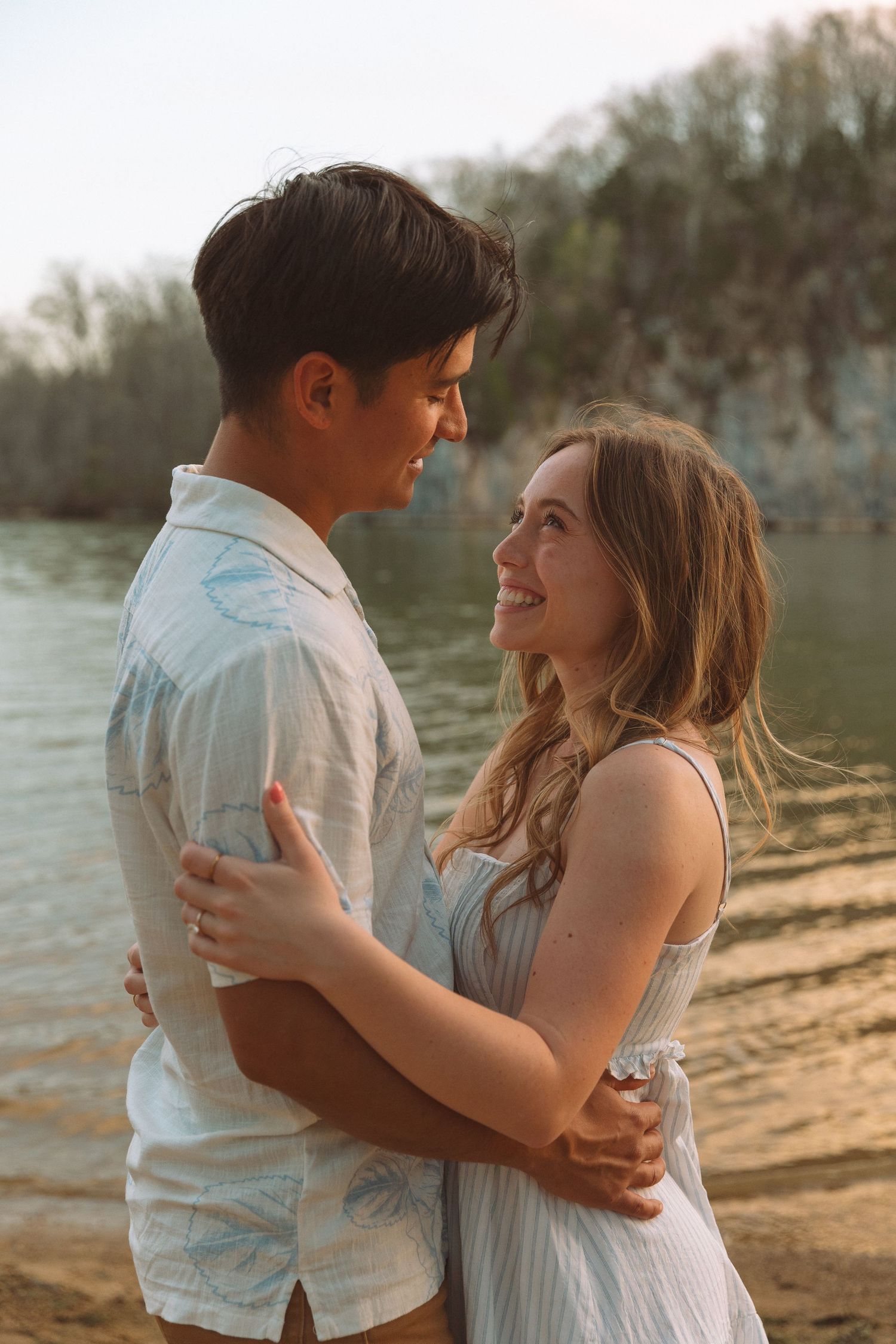 Smiling couple embraces lovingly by a river at sunset during engagement photography session.