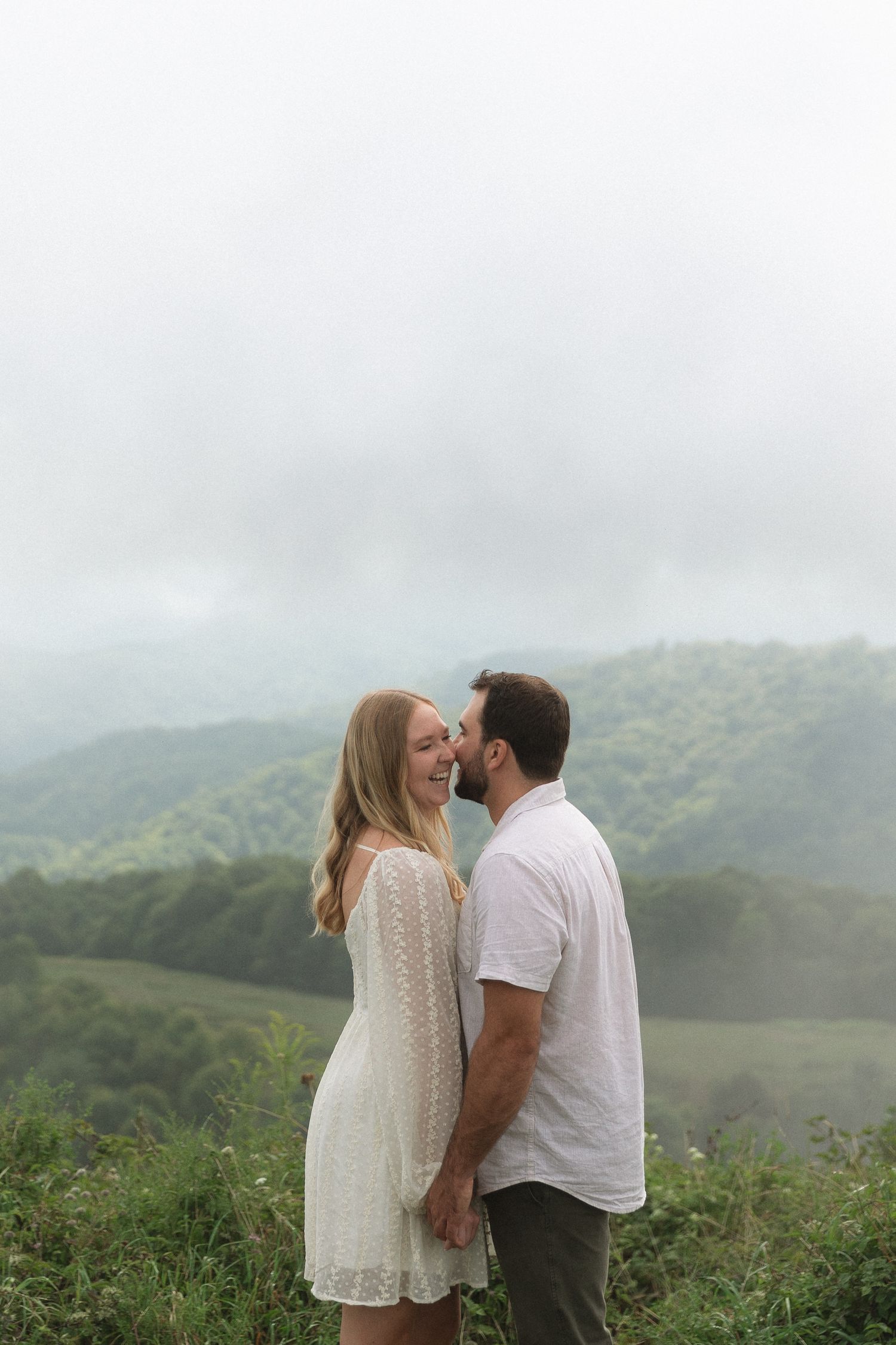 Happy couple laughing together on misty mountain overlook with lush green valley background.