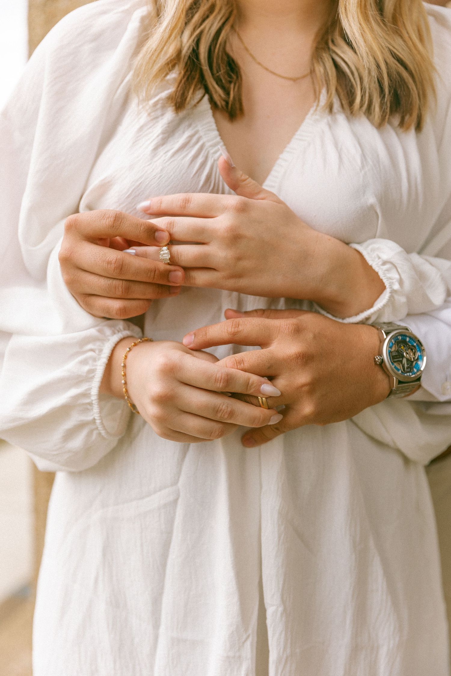 Close-up of couple holding hands, woman wearing diamond engagement ring and gold bracelet, man wearing a colorful watch.