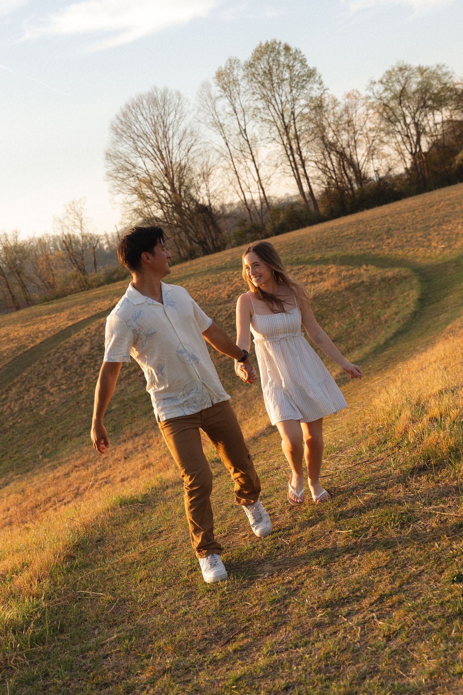 Young couple holds hands walking through sunlit golden field during sunset engagement session.