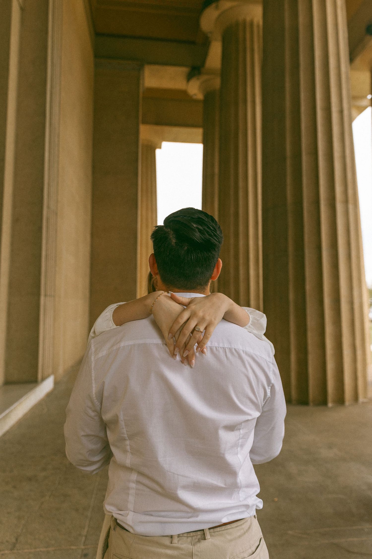 Couple embracing from behind near tall stone columns, woman's hand with engagement ring resting on man's shoulder.