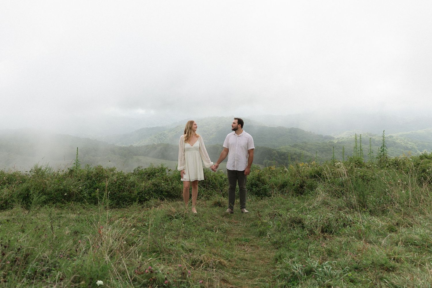 Couple holding hands on misty mountain hilltop, woman in white dress, lush green landscape with foggy mountains behind.