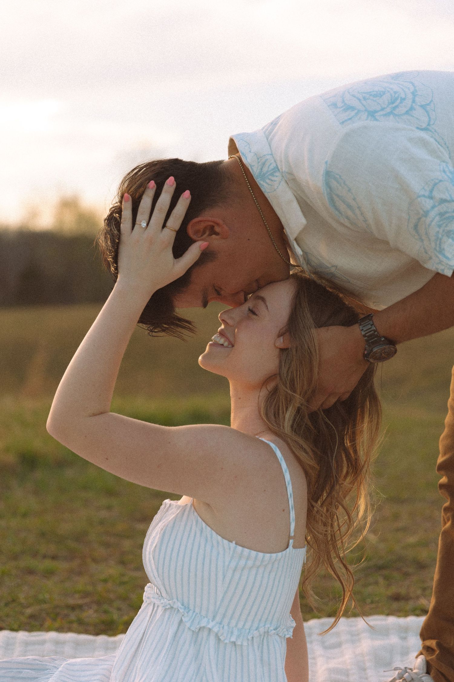 Joyful woman smiling as her partner leans down to kiss her forehead during a golden hour engagement session.