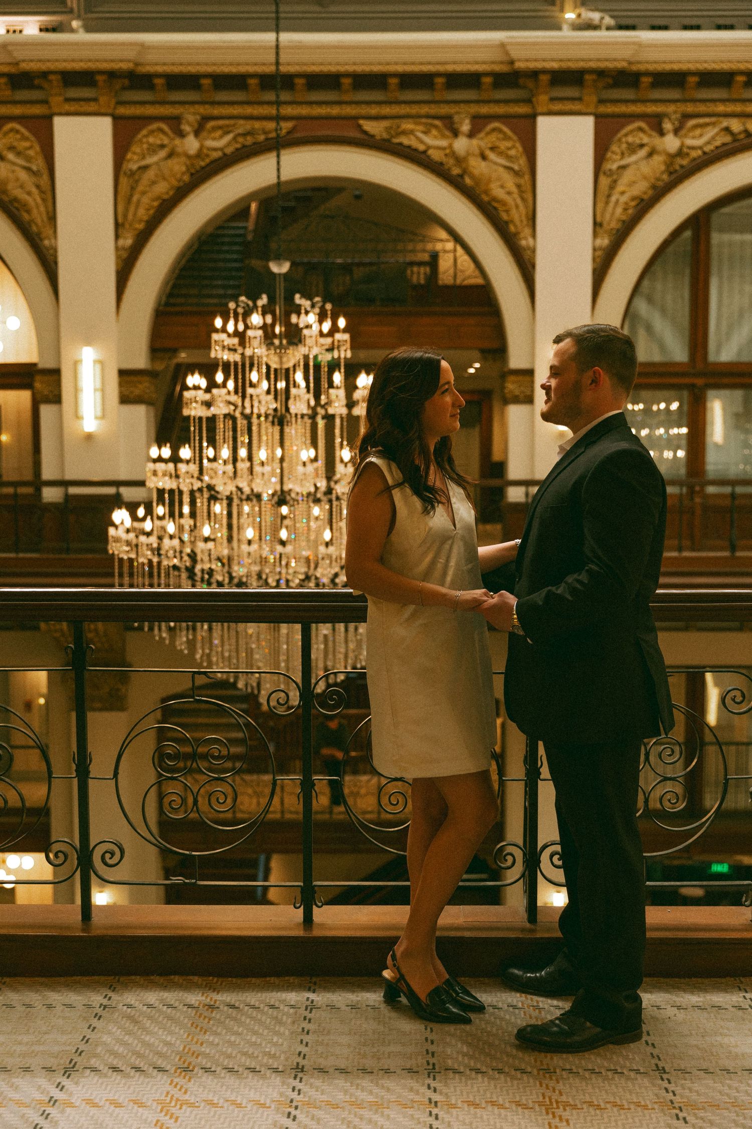 Couple holding hands on a balcony with ornate arches and a glowing chandelier behind them.
