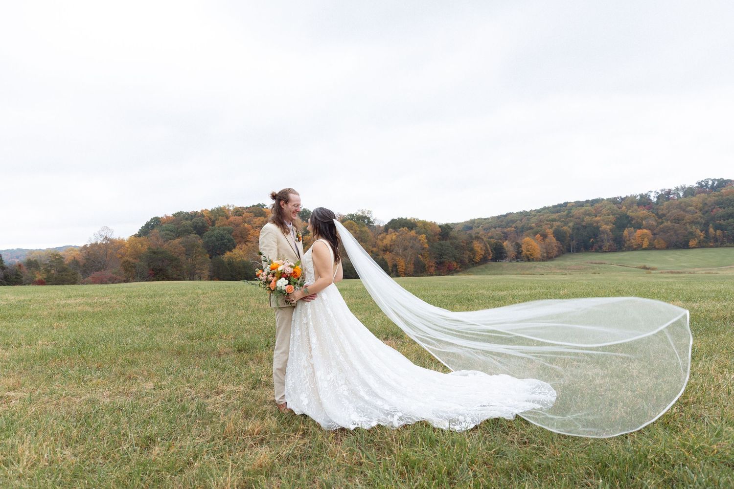 Bride and groom embrace in an open field with autumn foliage, bridal veil flowing in the wind.