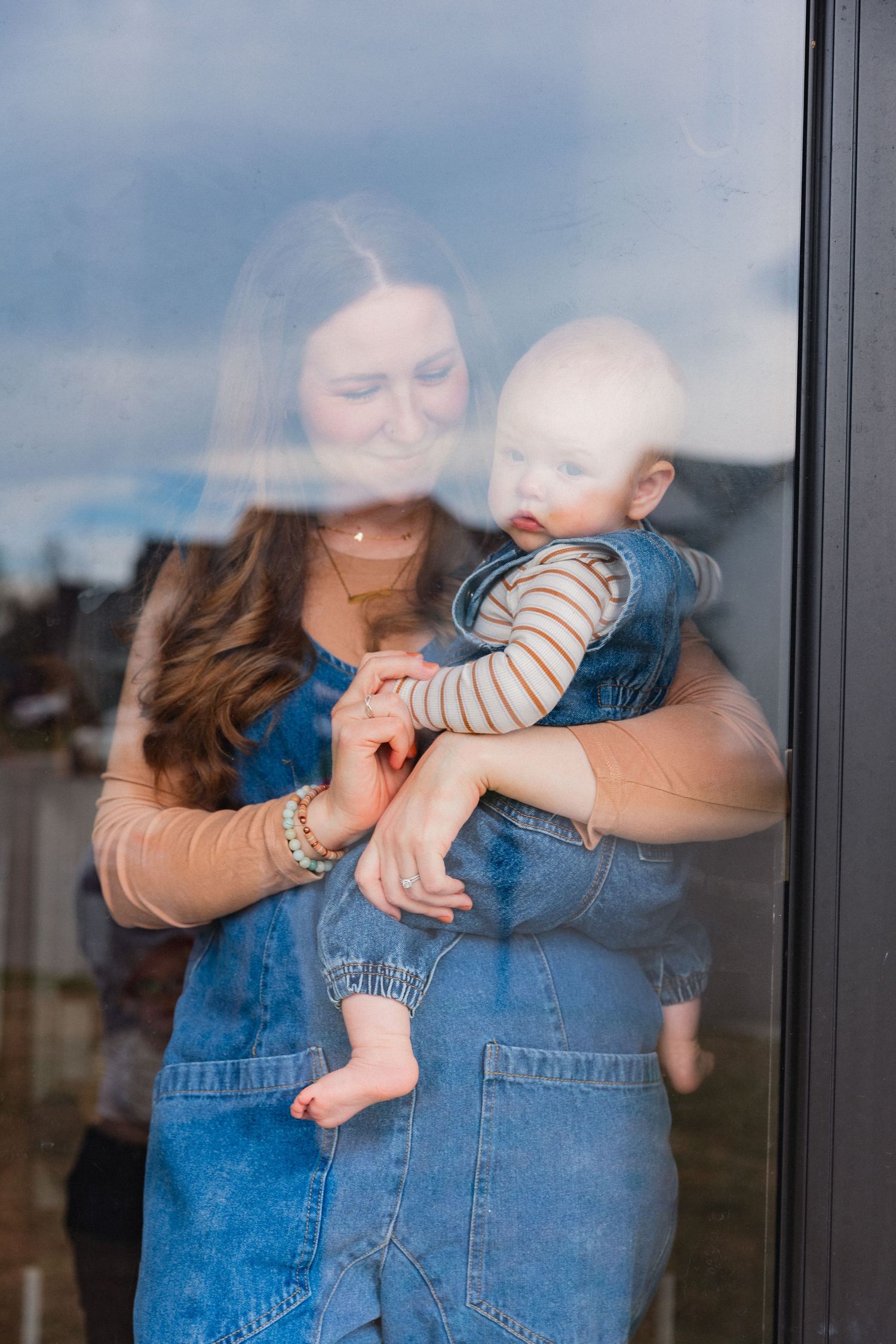 Mother holding baby infant viewed through a large window on a cloudy day.