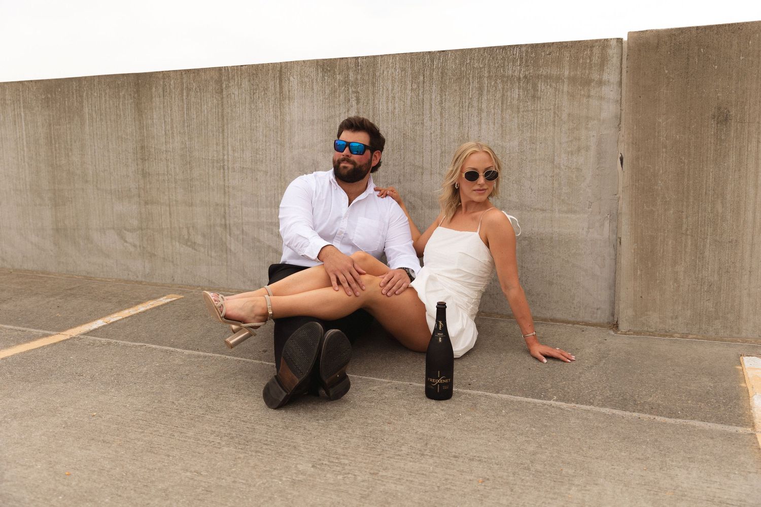 A stylish couple sitting against a concrete wall outdoors, wearing sunglasses, with a wine bottle placed beside them.