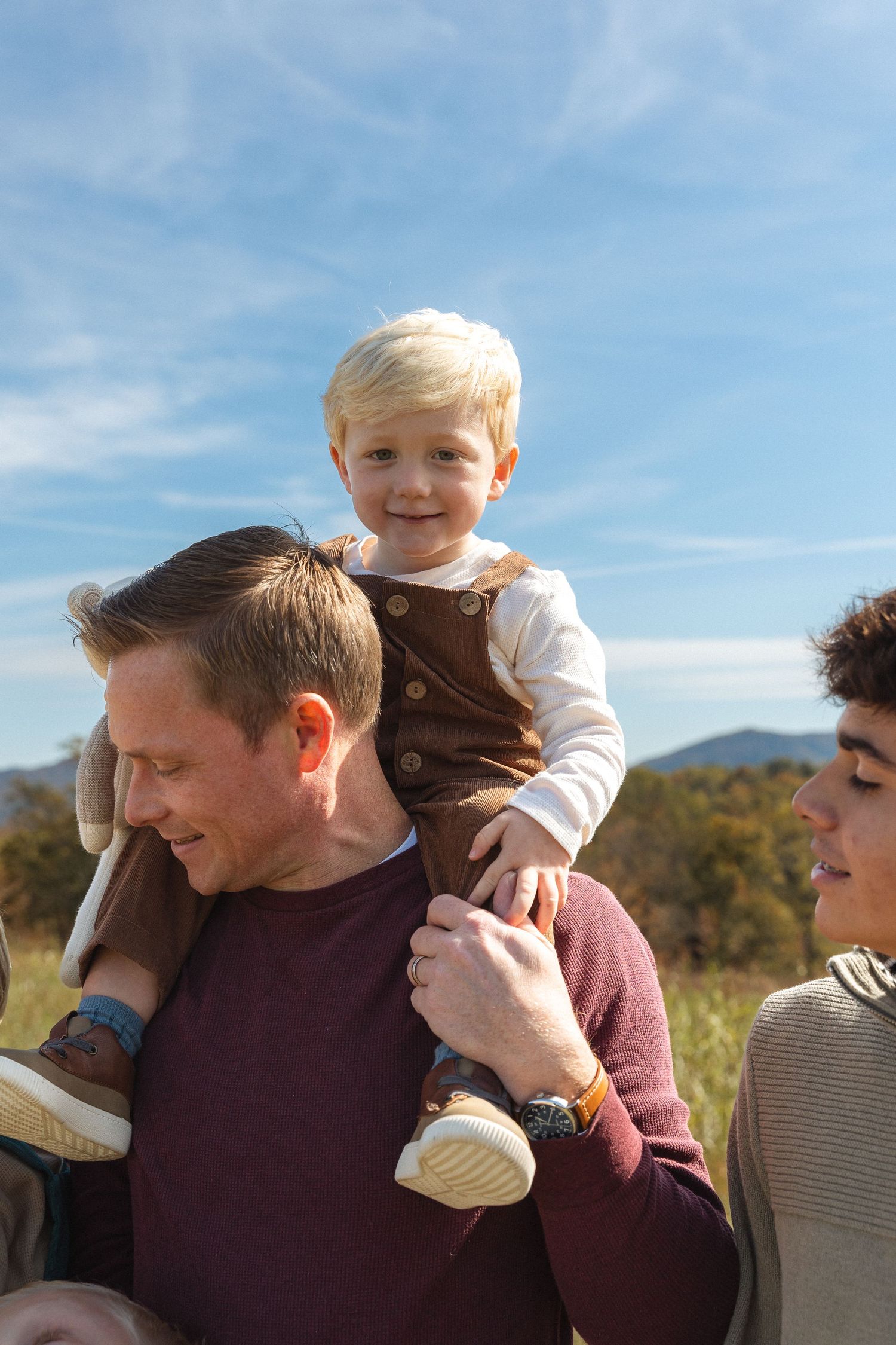 Happy toddler sitting on fathers shoulders smiling outdoors on a sunny day with mountains in background.