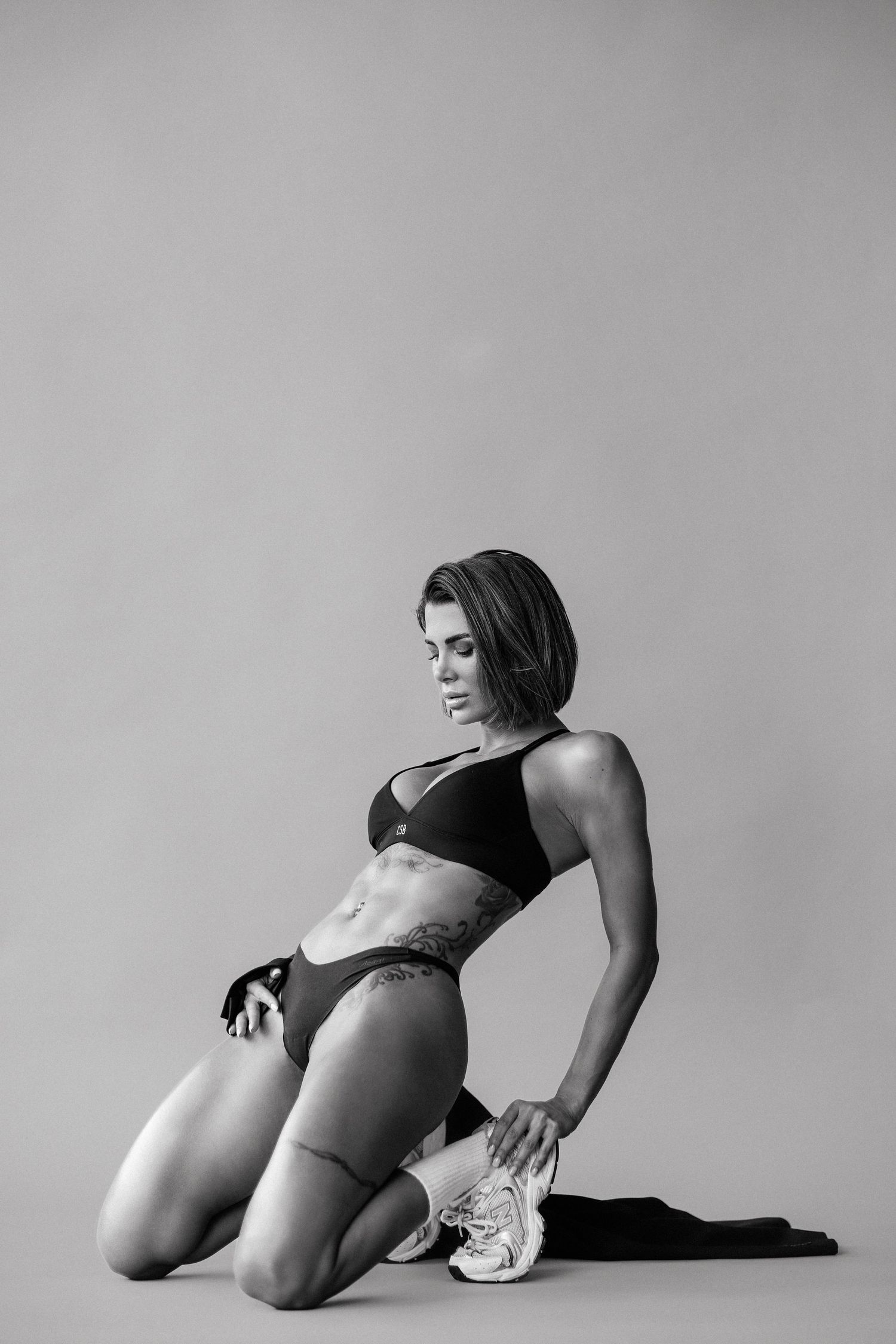 Fit woman in black sports bra and bikini bottom posing on knees in black and white studio photo.
