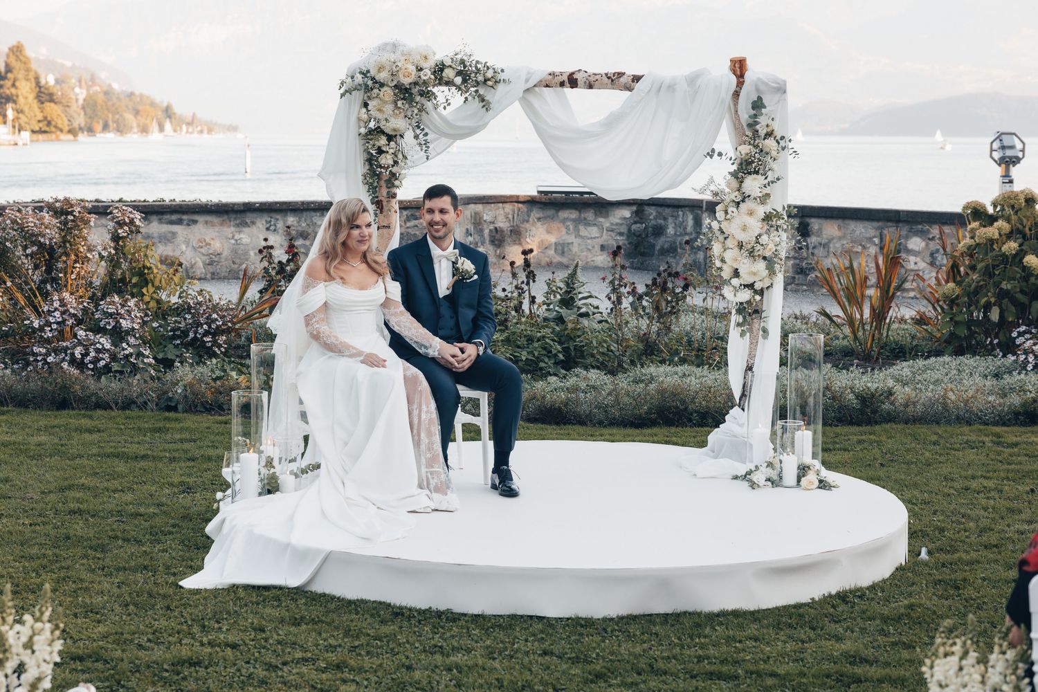 Bride and groom seated on white circular outdoor wedding altar with floral arch and waterfront backdrop.