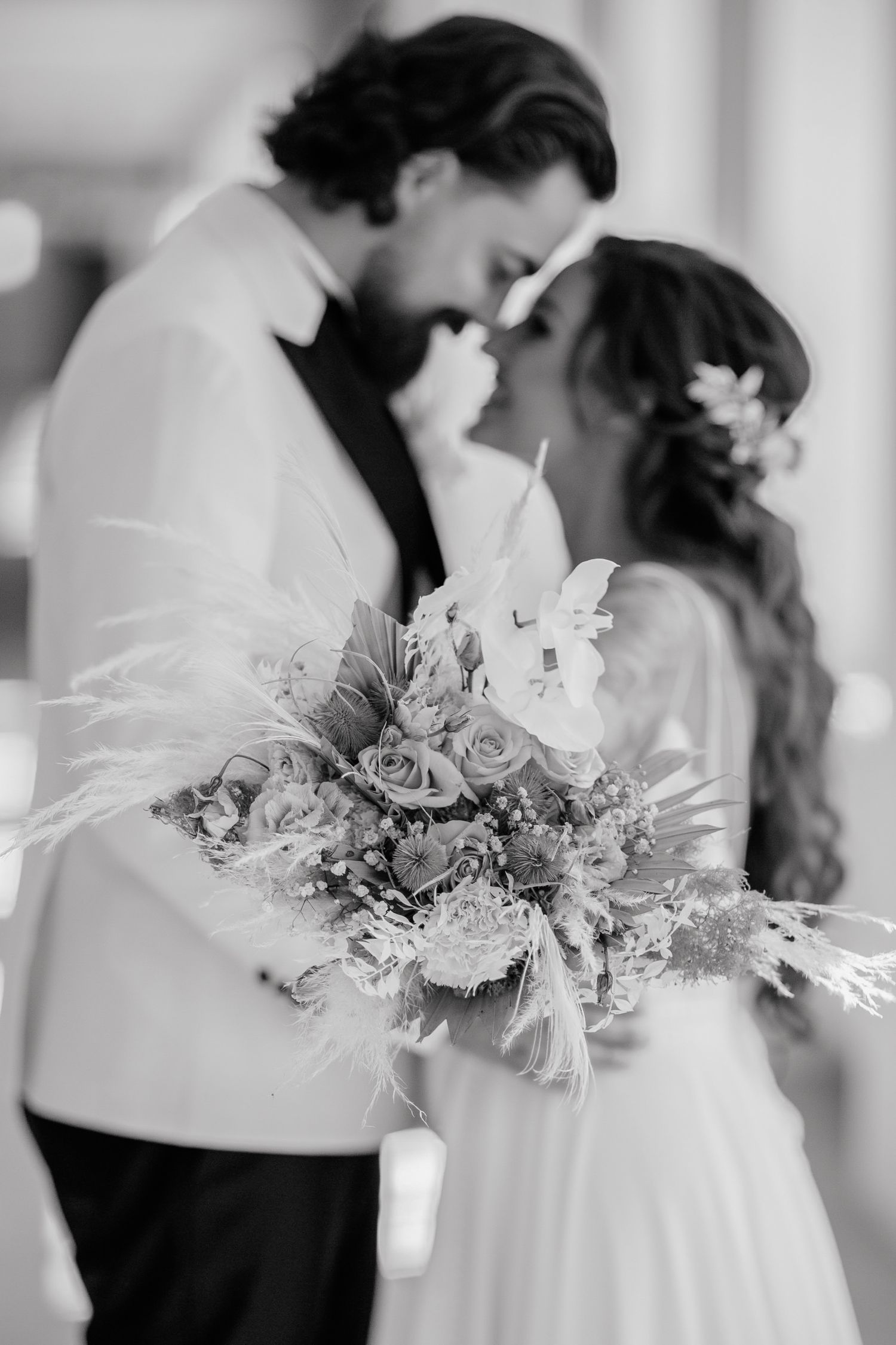 Black and white photo of a bride and groom almost kissing, with a lush floral bouquet featuring roses and feathers.