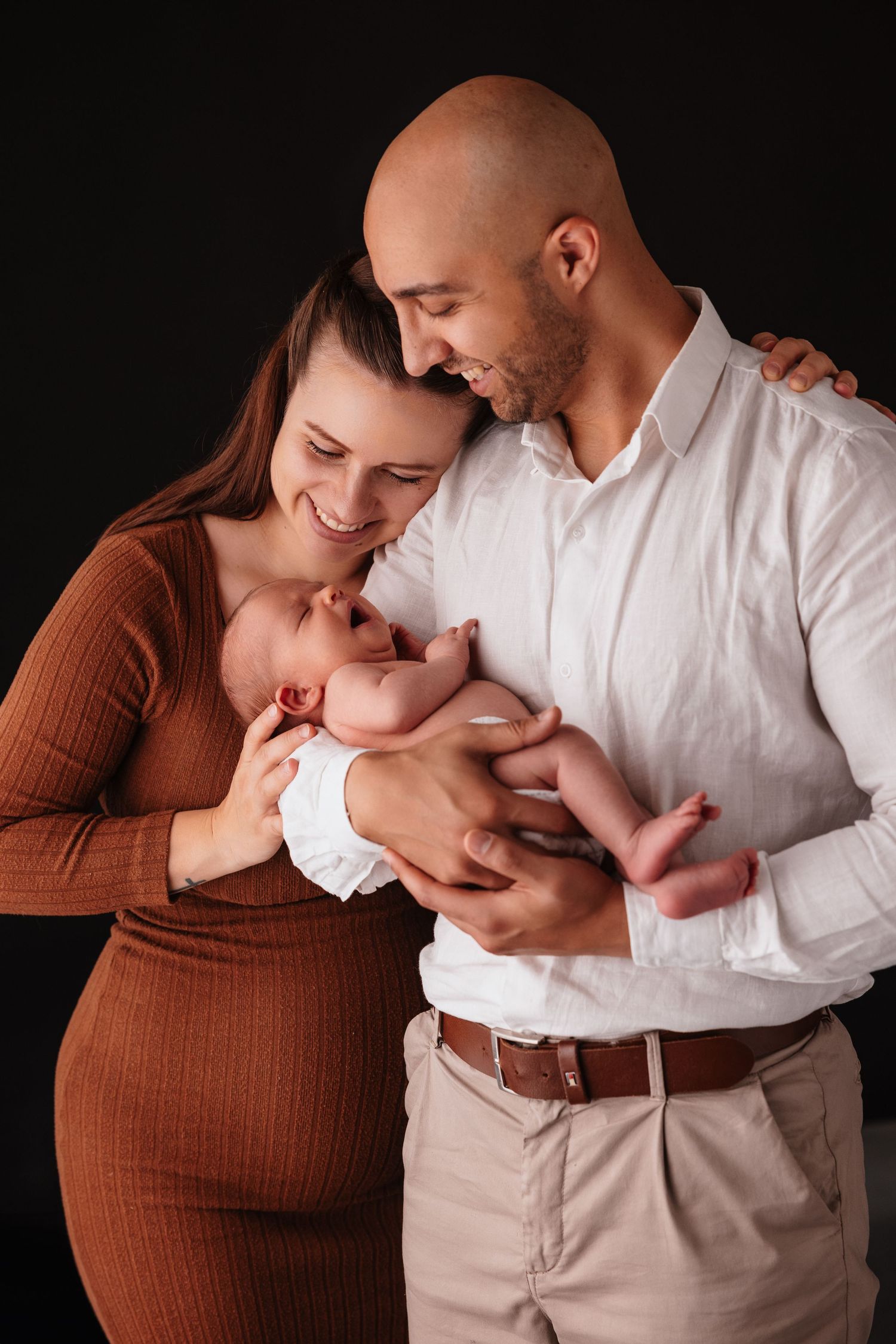 A couple smiles lovingly at their newborn baby in a professional studio portrait against a dark background.