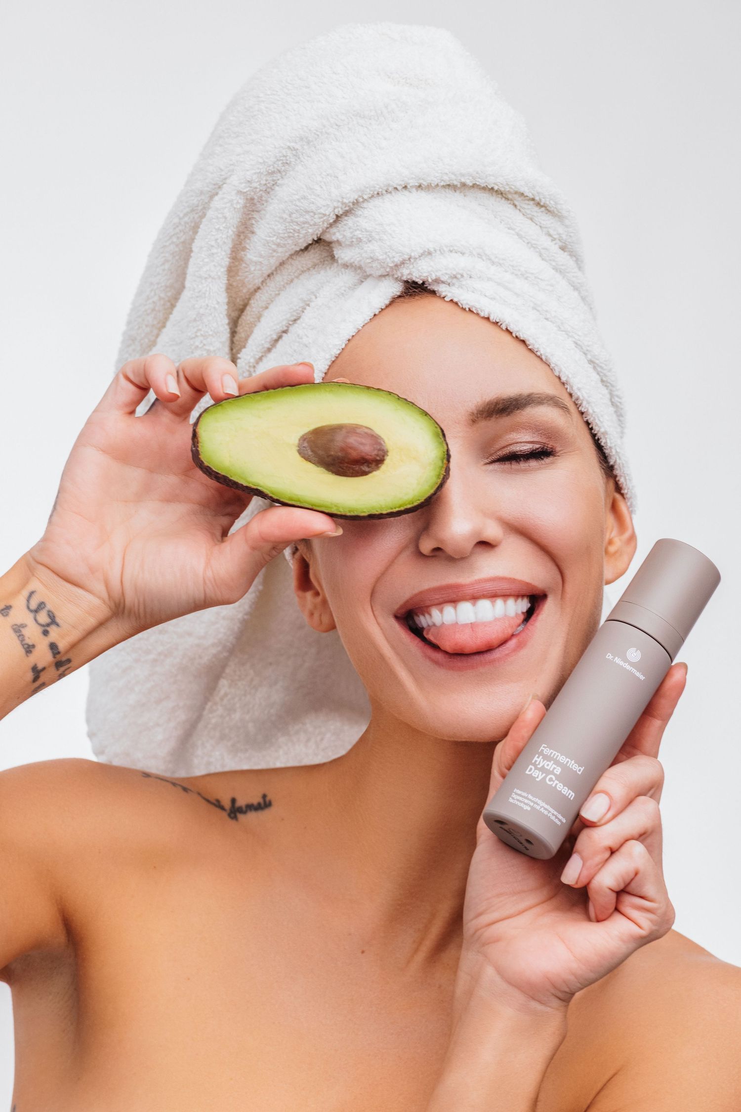 Smiling woman with towel on head holds avocado half over eye and skincare product bottle against white background.