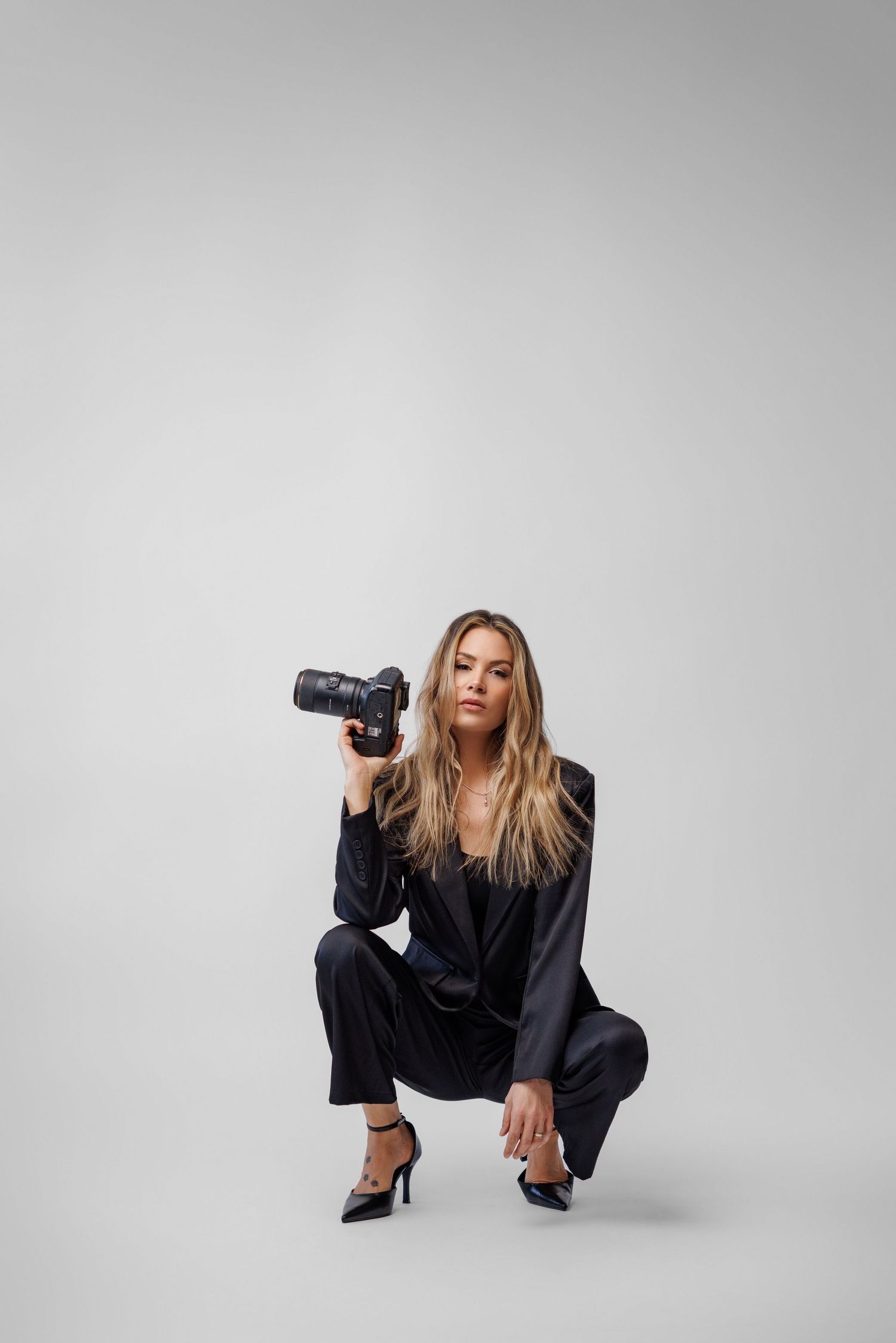 Blonde woman in dark suit crouching while holding a professional camera, studio photography portrait on gray background.