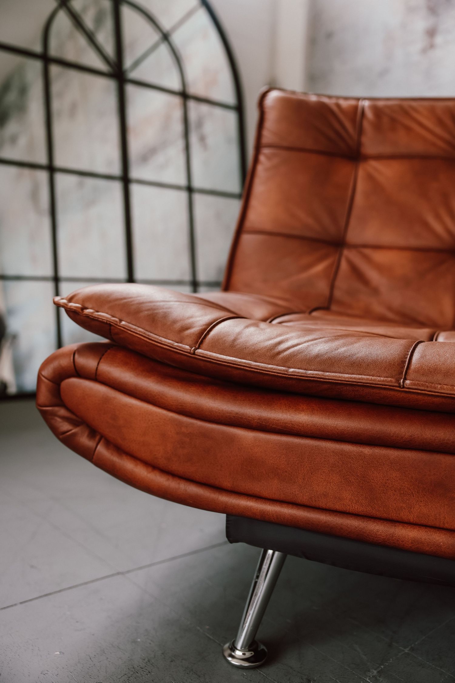 Vintage brown leather tufted armchair with chrome legs on gray floor near arched window.