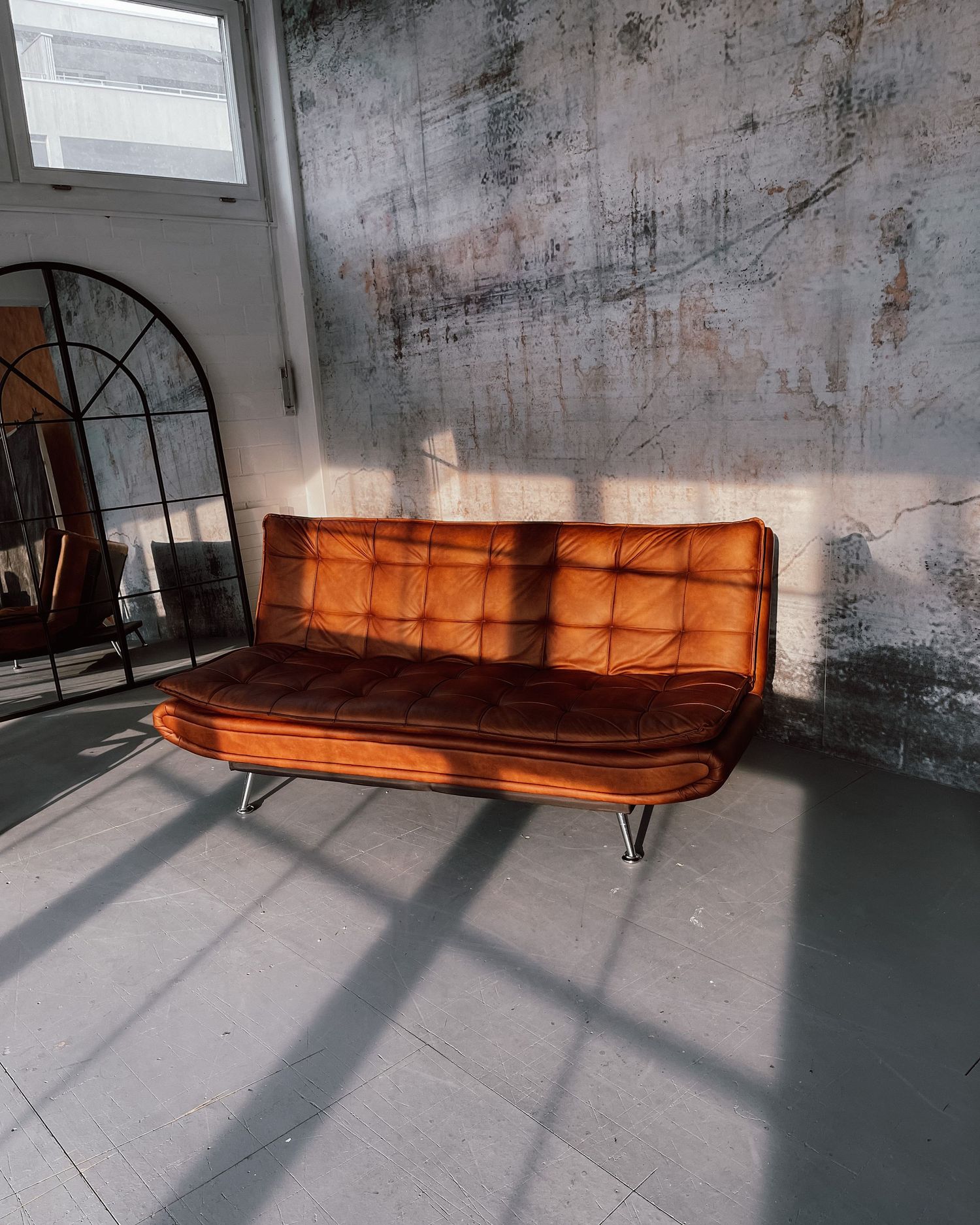 Vintage orange tufted velvet sofa with chrome legs in sunlit industrial loft space with distressed walls.