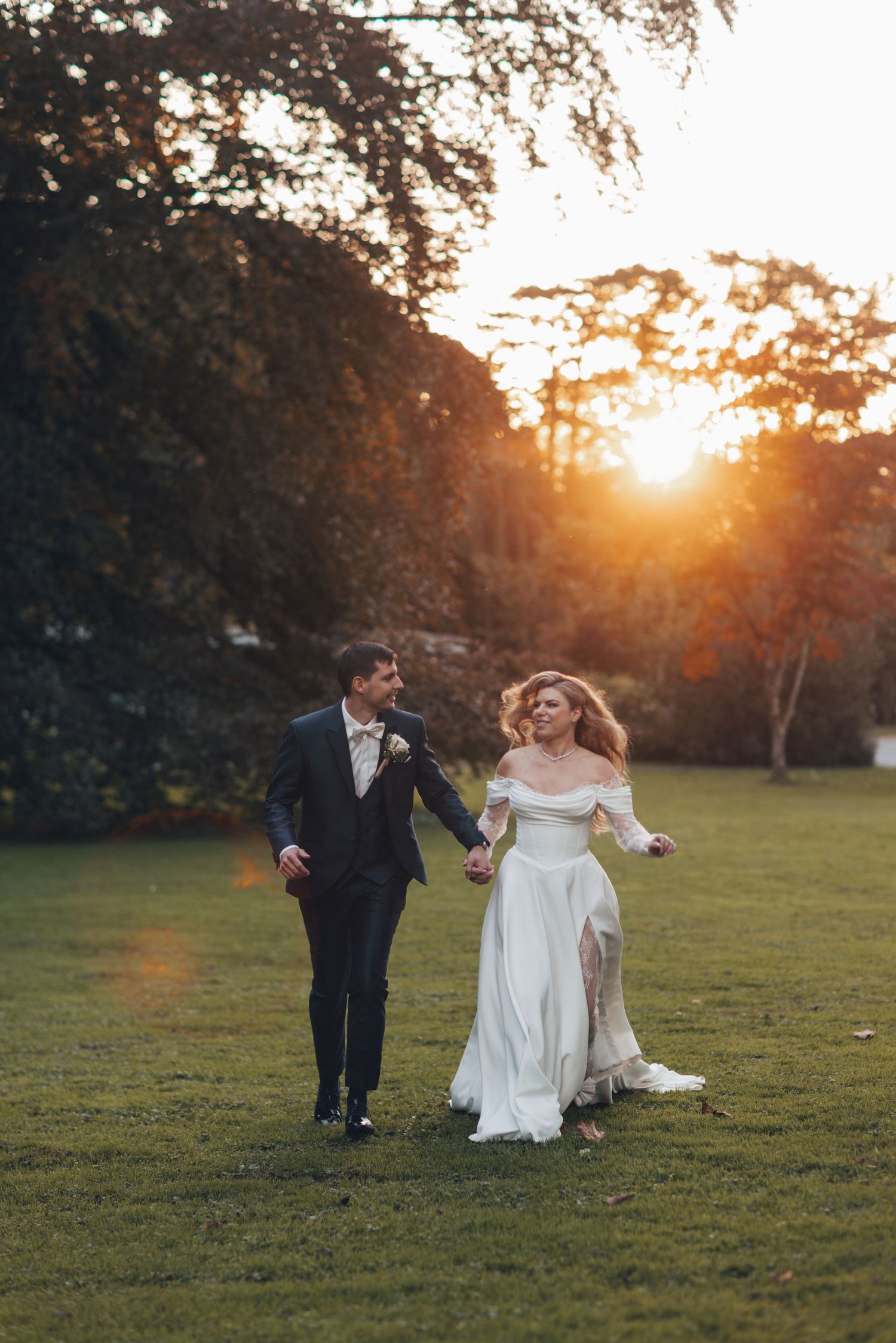 A bride and groom walk hand-in-hand across a green lawn at golden sunset, surrounded by lush trees.