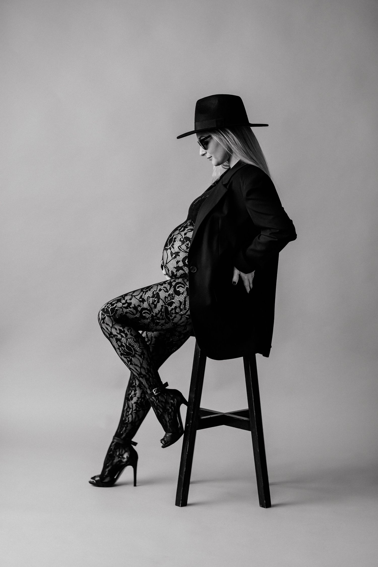 Pregnant woman in black blazer, lace tights, heels, and wide-brim hat poses on stool in black and white photo.