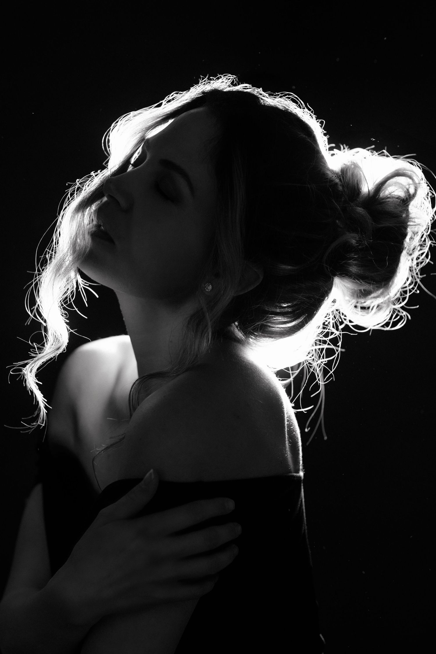 Black and white portrait of a woman with elegant updo hairstyle, backlit with dramatic rim lighting on dark background.