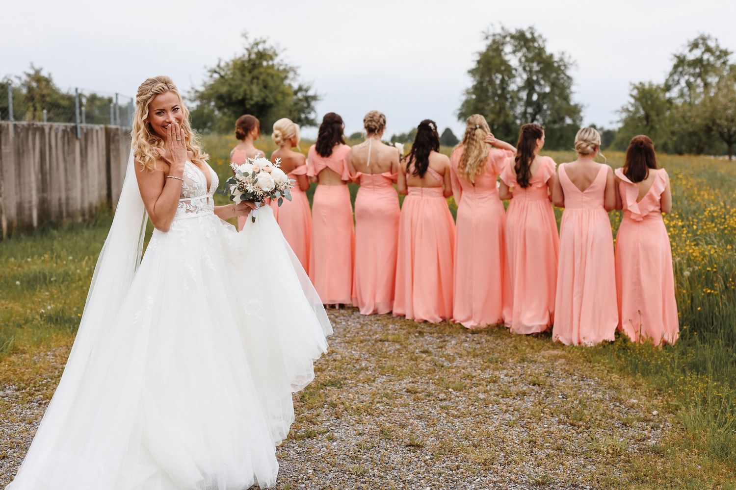 Bride in white gown holds bouquet while bridesmaids in coral dresses pose with backs turned on a gravel path.