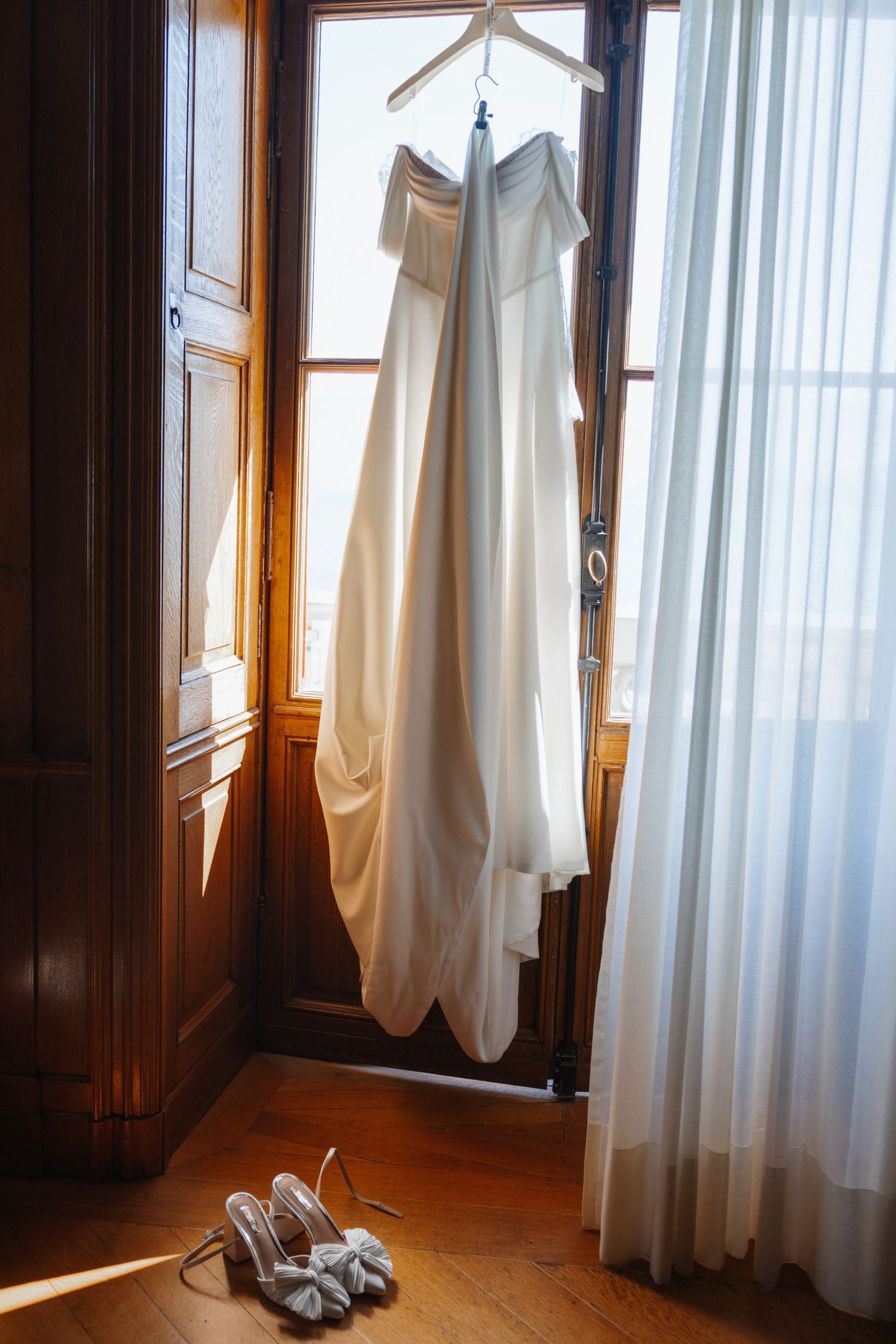 White wedding dress hanging on door with silver heeled shoes on wooden floor below, soft natural light.