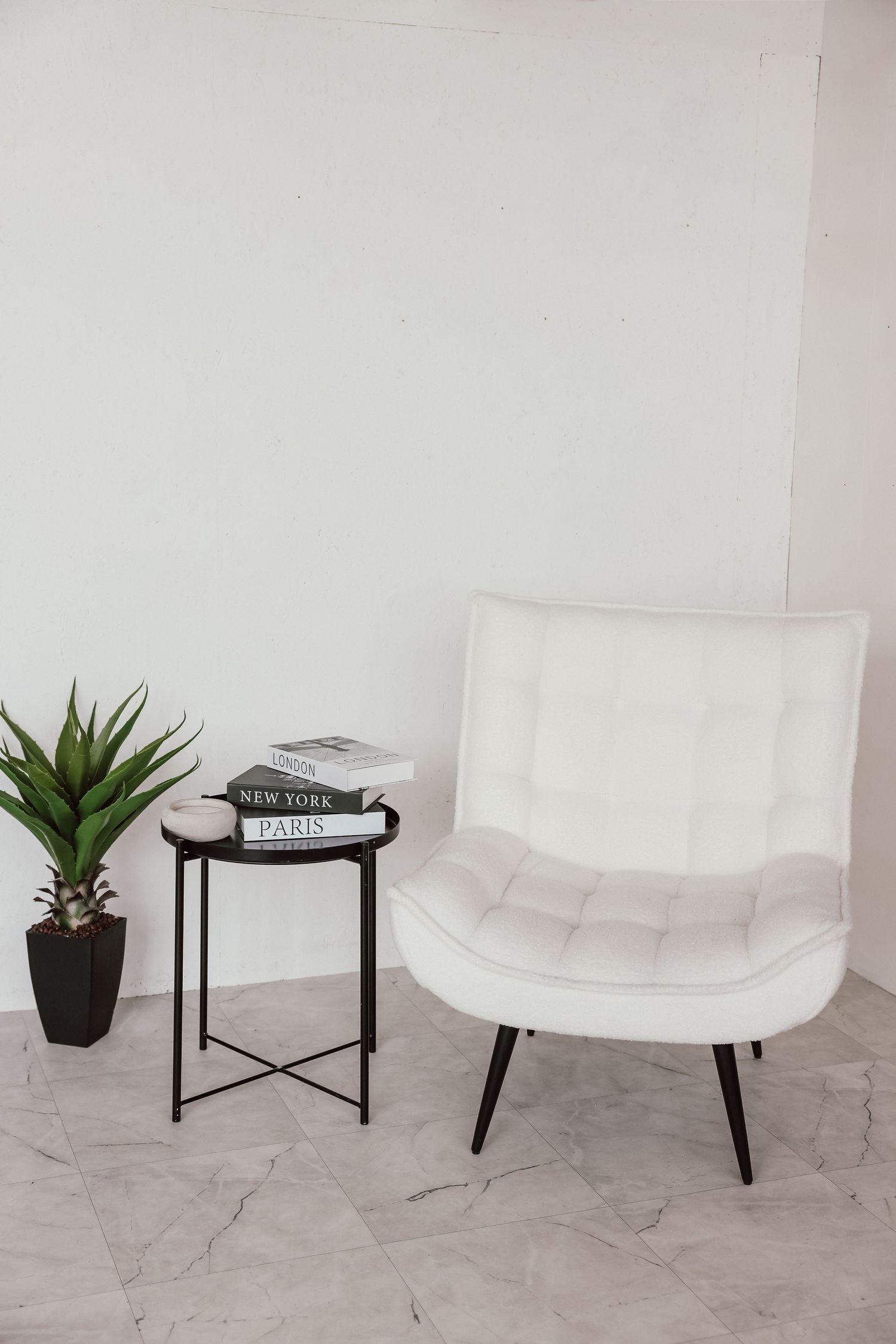 White tufted accent chair beside black metal side table with books and plant on marble floor against white wall.