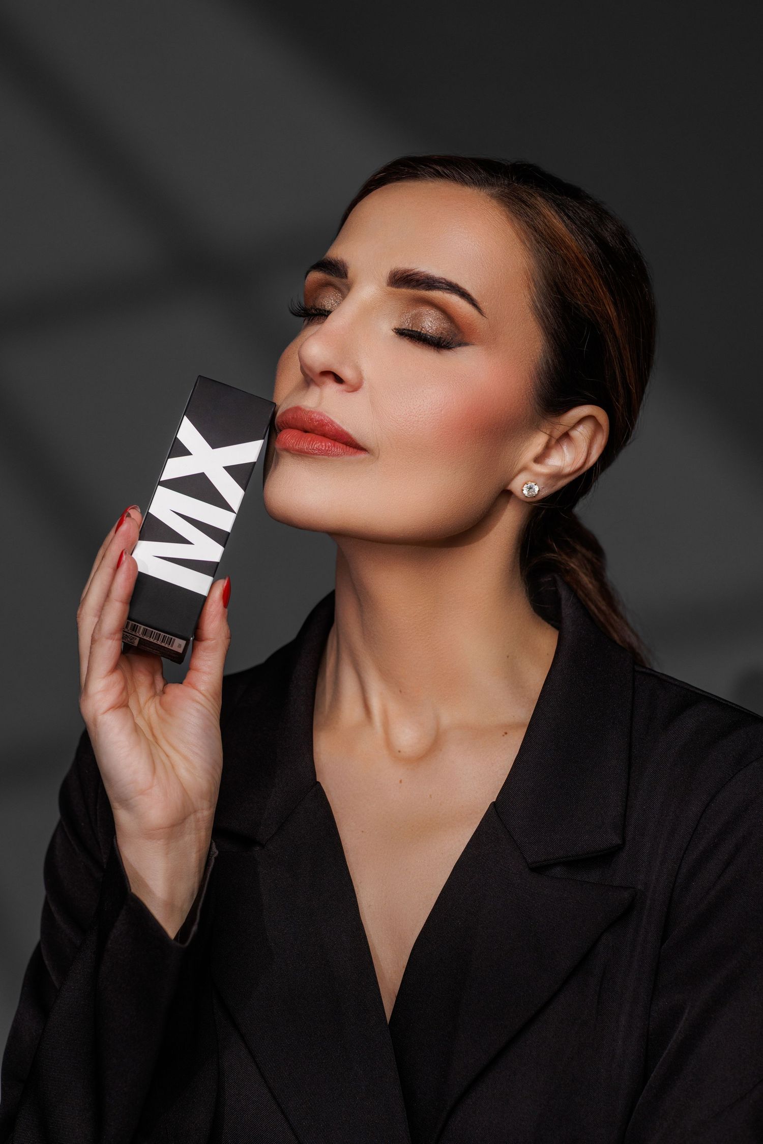 Elegant woman in black blazer holding MX branded beauty product box against gray background.
