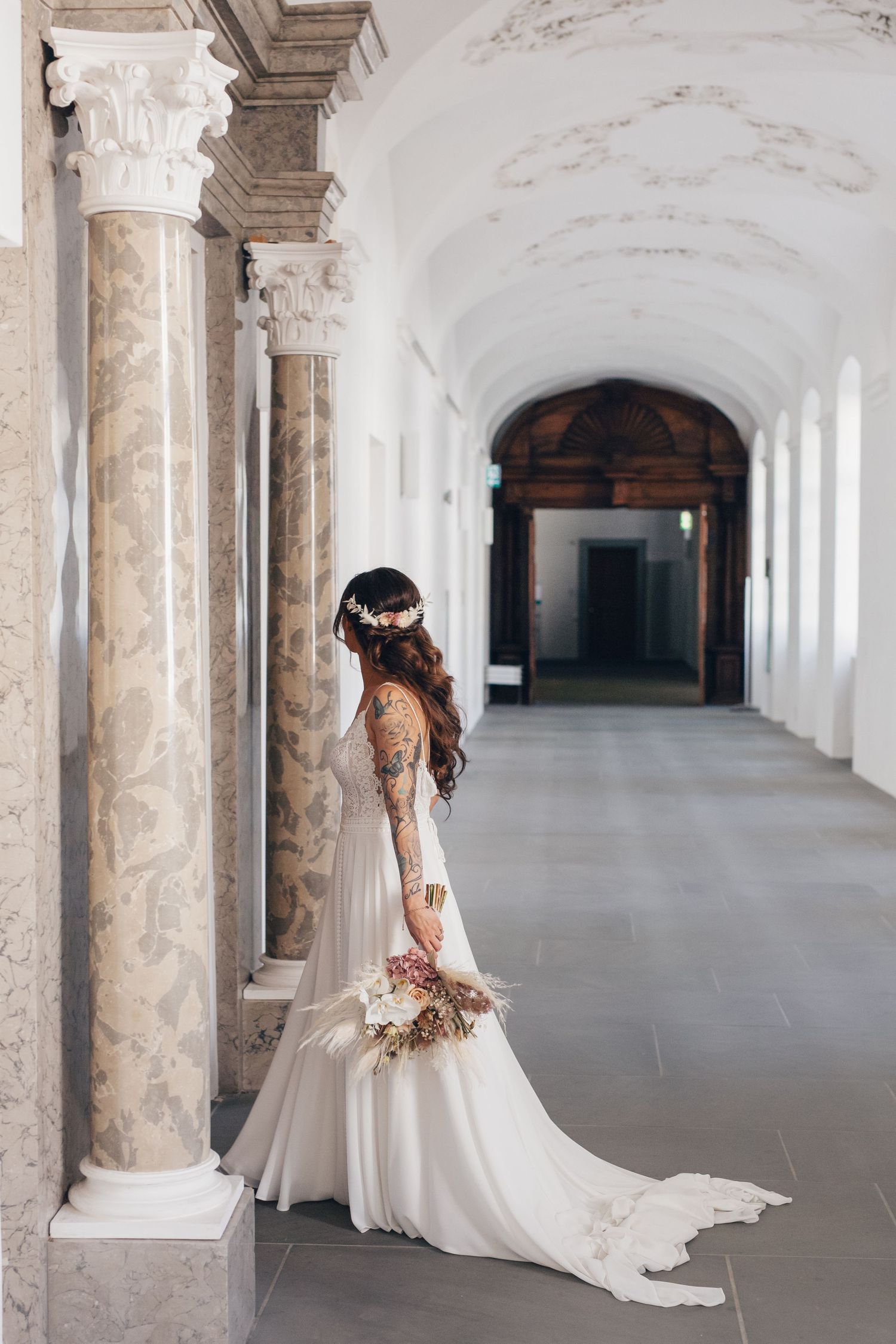 Bride in white lace gown with tattoos holds bouquet beside marble columns in elegant arched hallway.