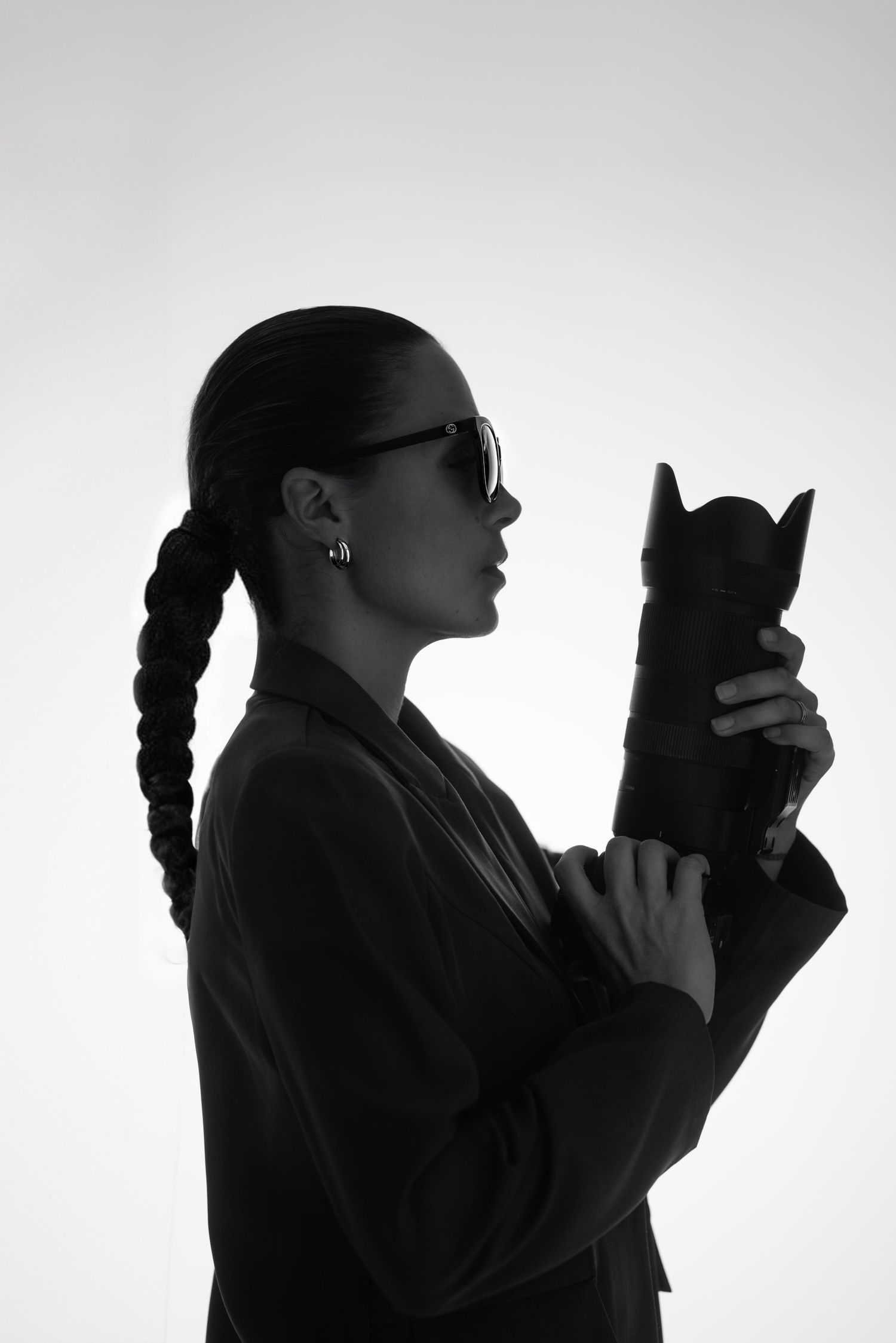 Black and white side profile silhouette of a woman in sunglasses and a blazer with a long braid, holding a large camera lens.