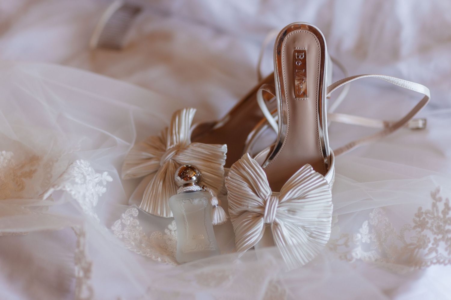 Elegant white bridal heels with bow detail beside a perfume bottle on lace wedding veil.