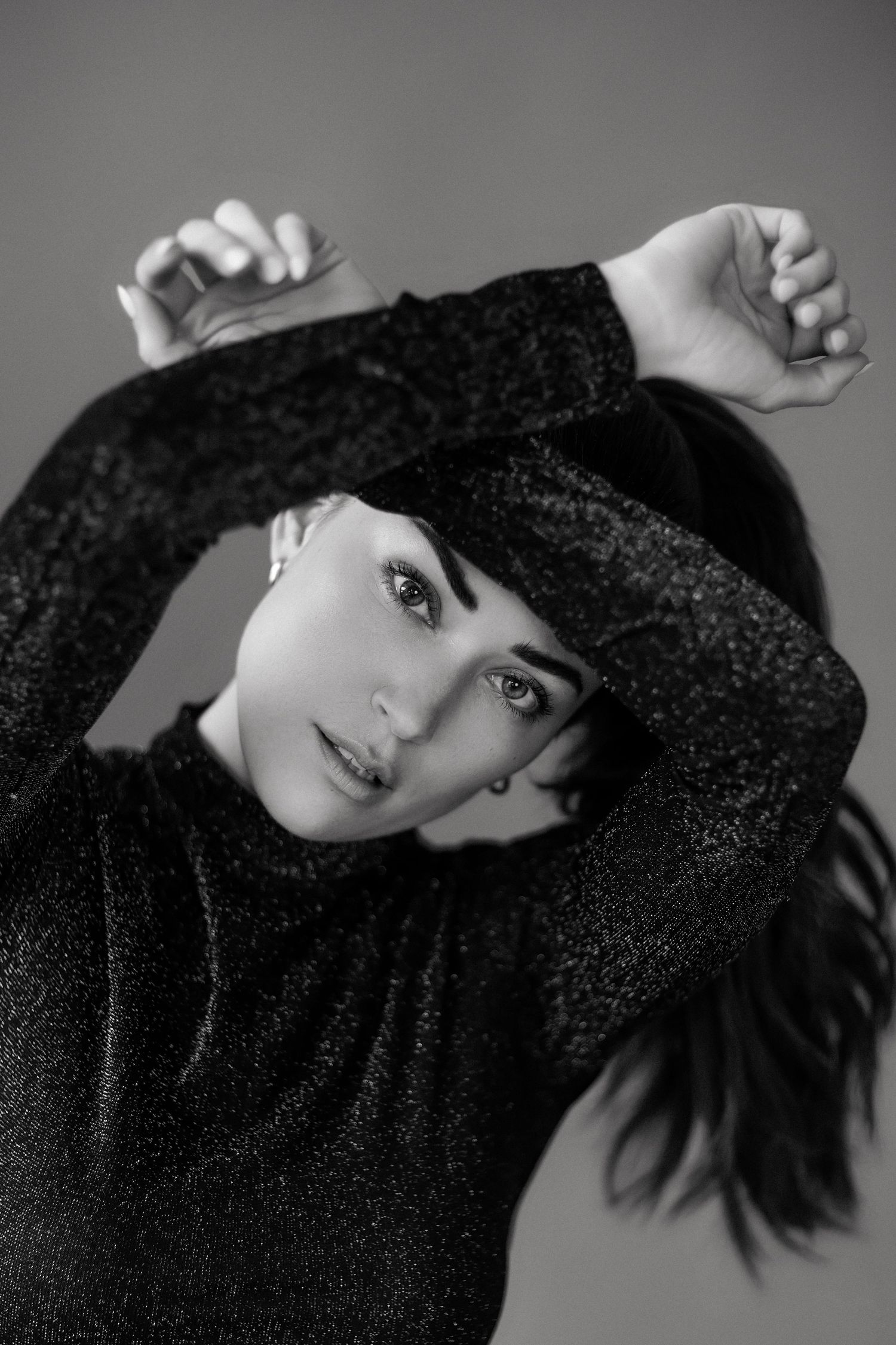 Black and white portrait of a young woman in a sparkly dark top, arms raised gracefully above her head.