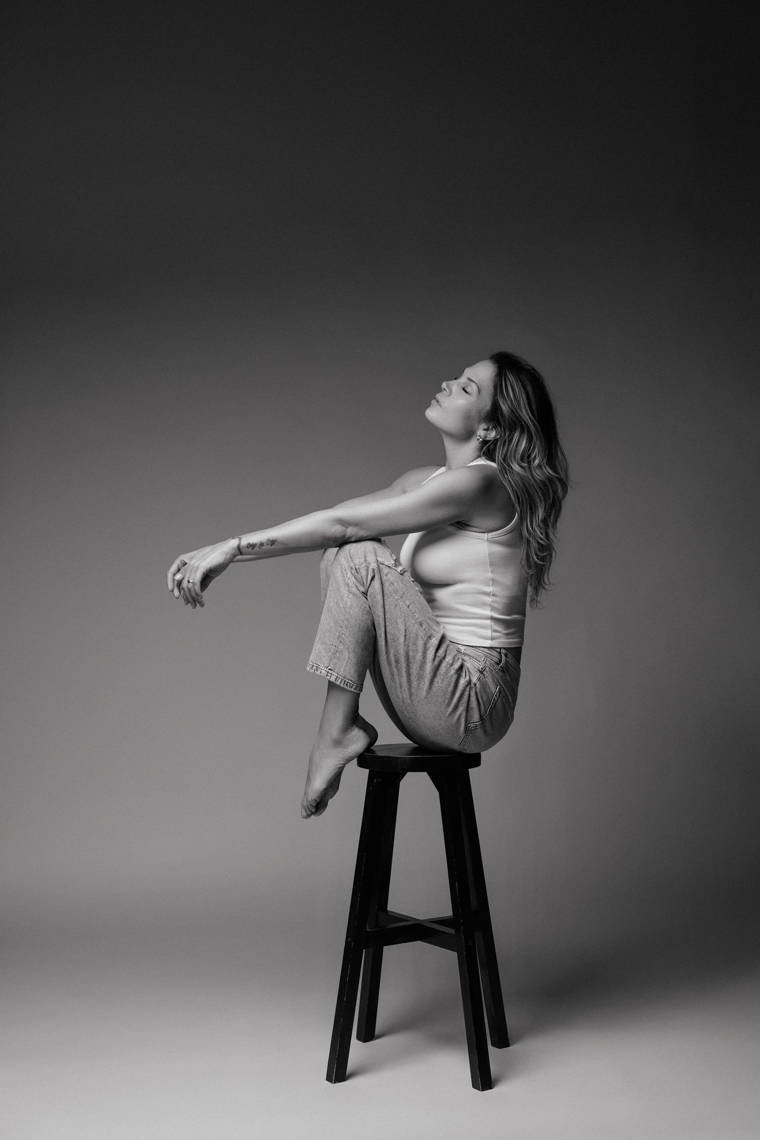 Black and white studio portrait of a woman in a white tank top and light pants sitting on a tall dark stool with her head tilted back.