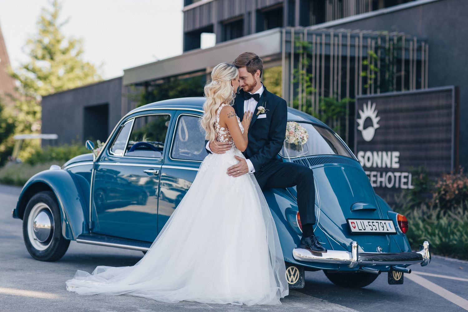 Bride and groom kissing beside a vintage blue VW Beetle outside Sonne Hotel on their wedding day.