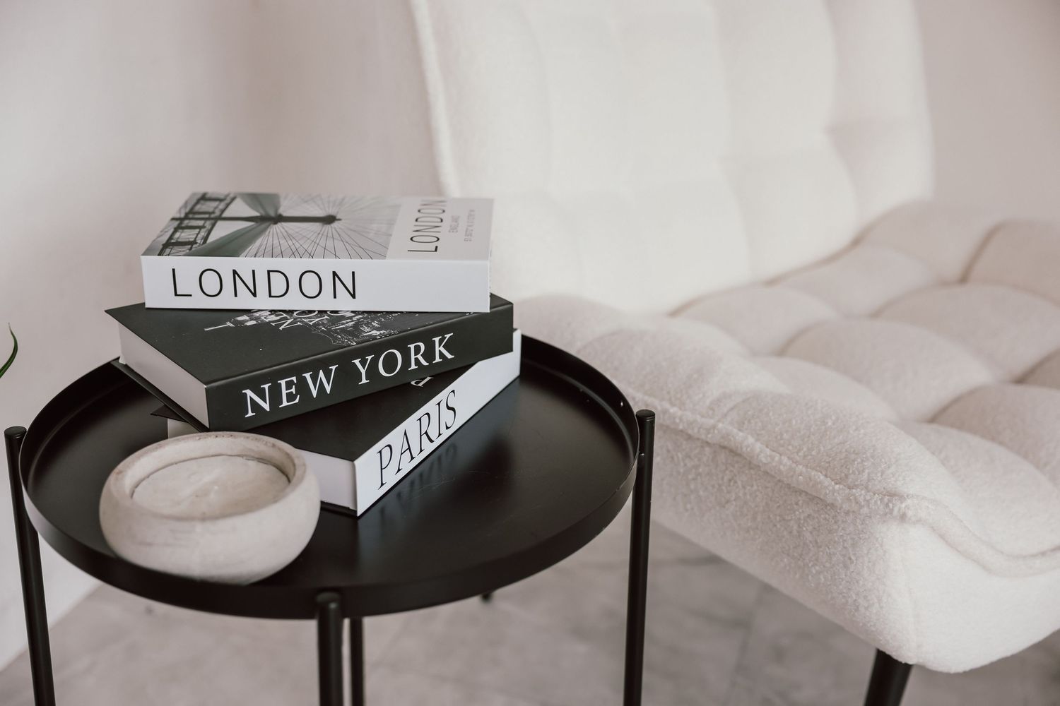 Black side table with London, New York, and Paris books stacked beside a white boucle chair in a minimalist room.