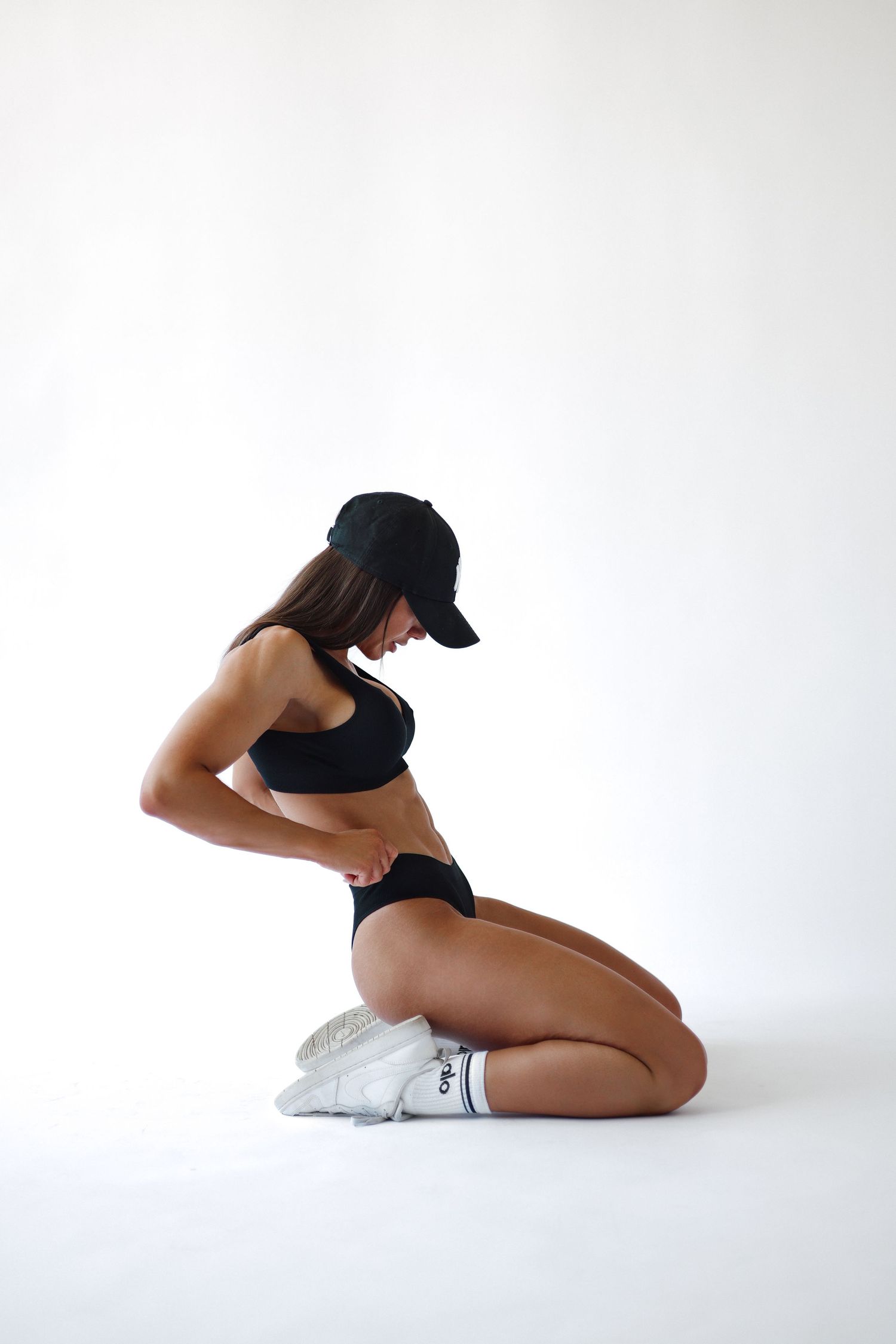 Fit woman in black sports bra, cap, and white socks kneeling on white background in athletic pose.
