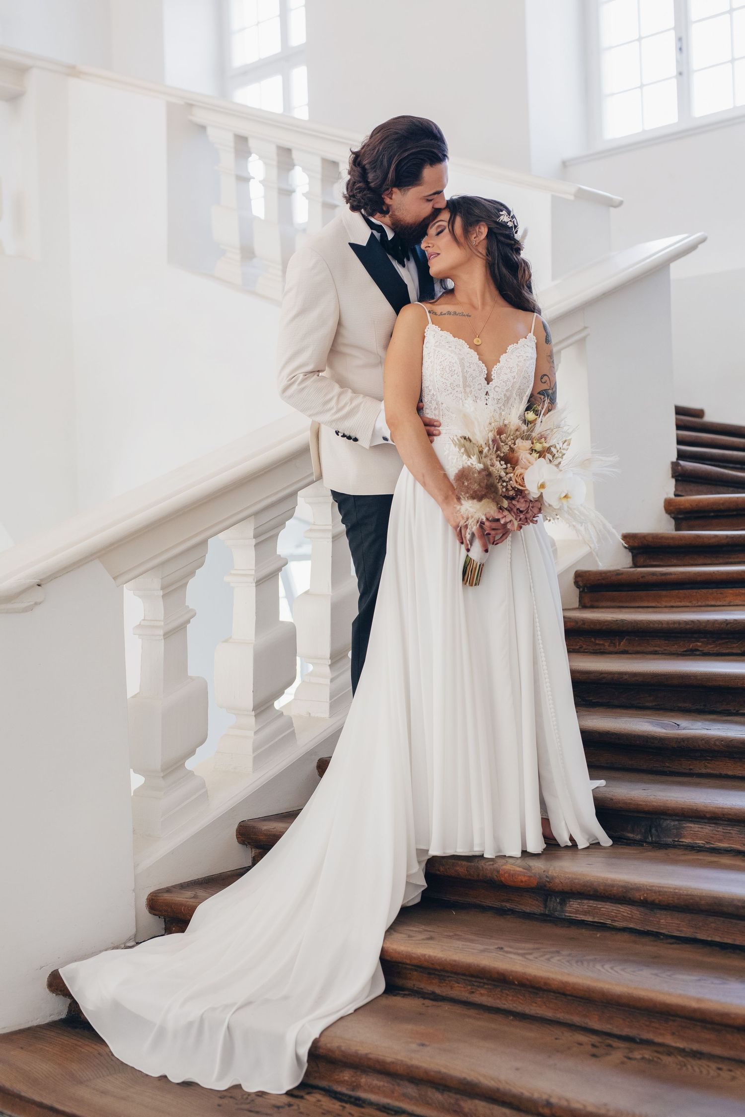 Bride and groom pose romantically on elegant white staircase, bride holding boho bouquet in flowing white gown.