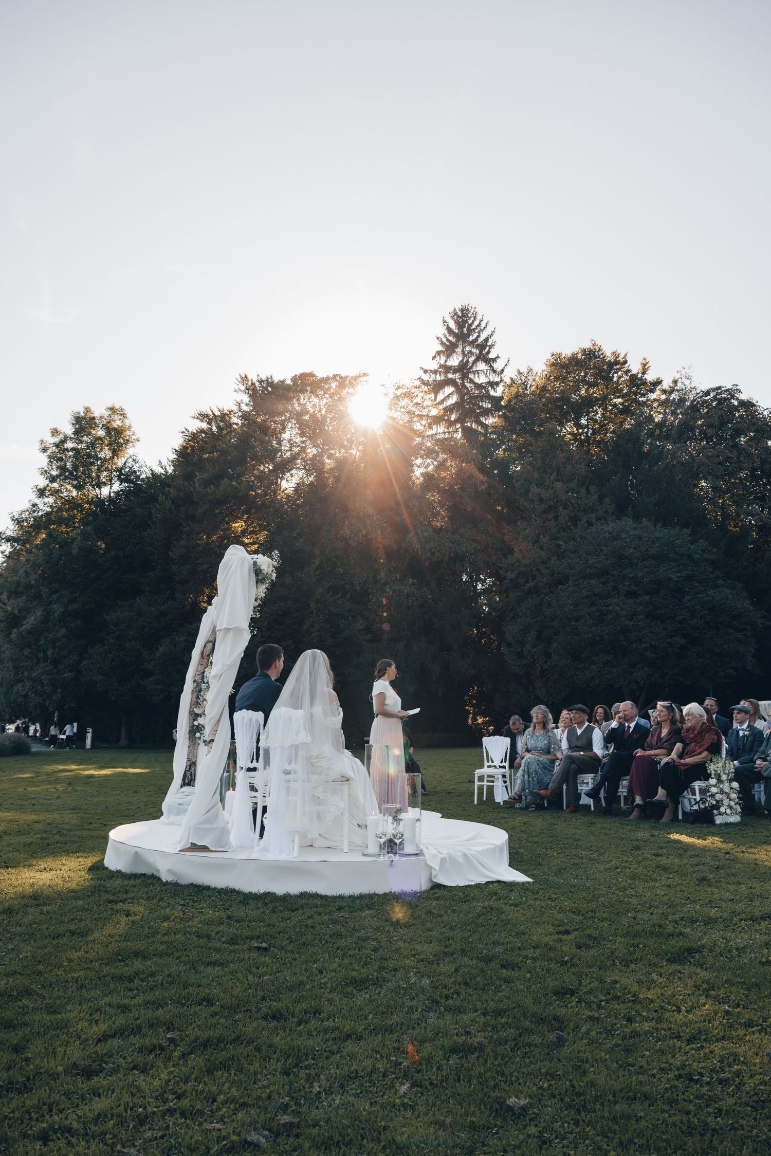 Outdoor wedding ceremony at sunset on white circular platform with bride, groom, and officiant, guests seated on lawn.