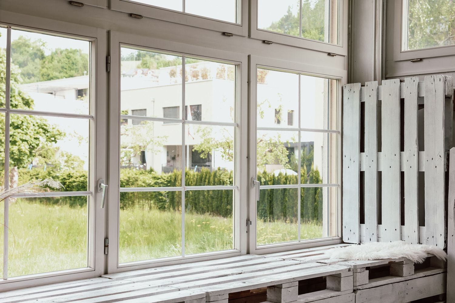 White wooden bench and pallet furniture by large bright windows overlooking a lush green garden.