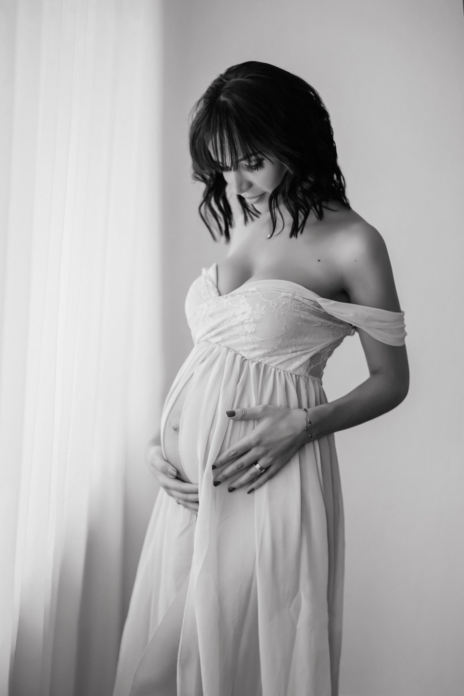 Black and white maternity photo of a pregnant woman in white off-shoulder dress cradling her baby bump.
