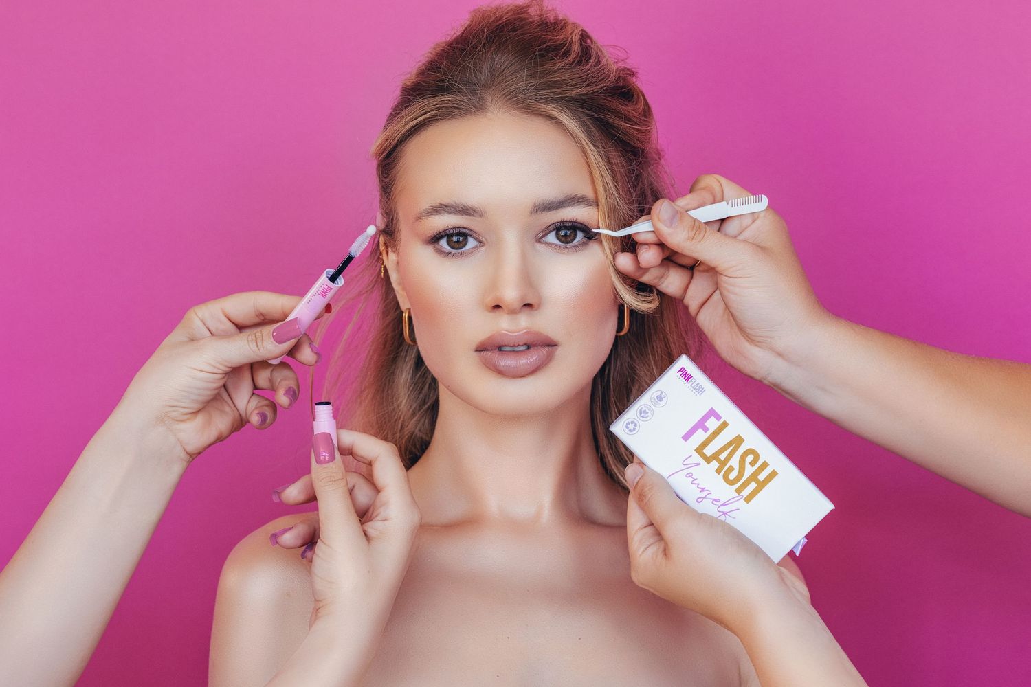 Young woman with blue eyes surrounded by hands applying mascara and holding Flash lash serum on pink background.