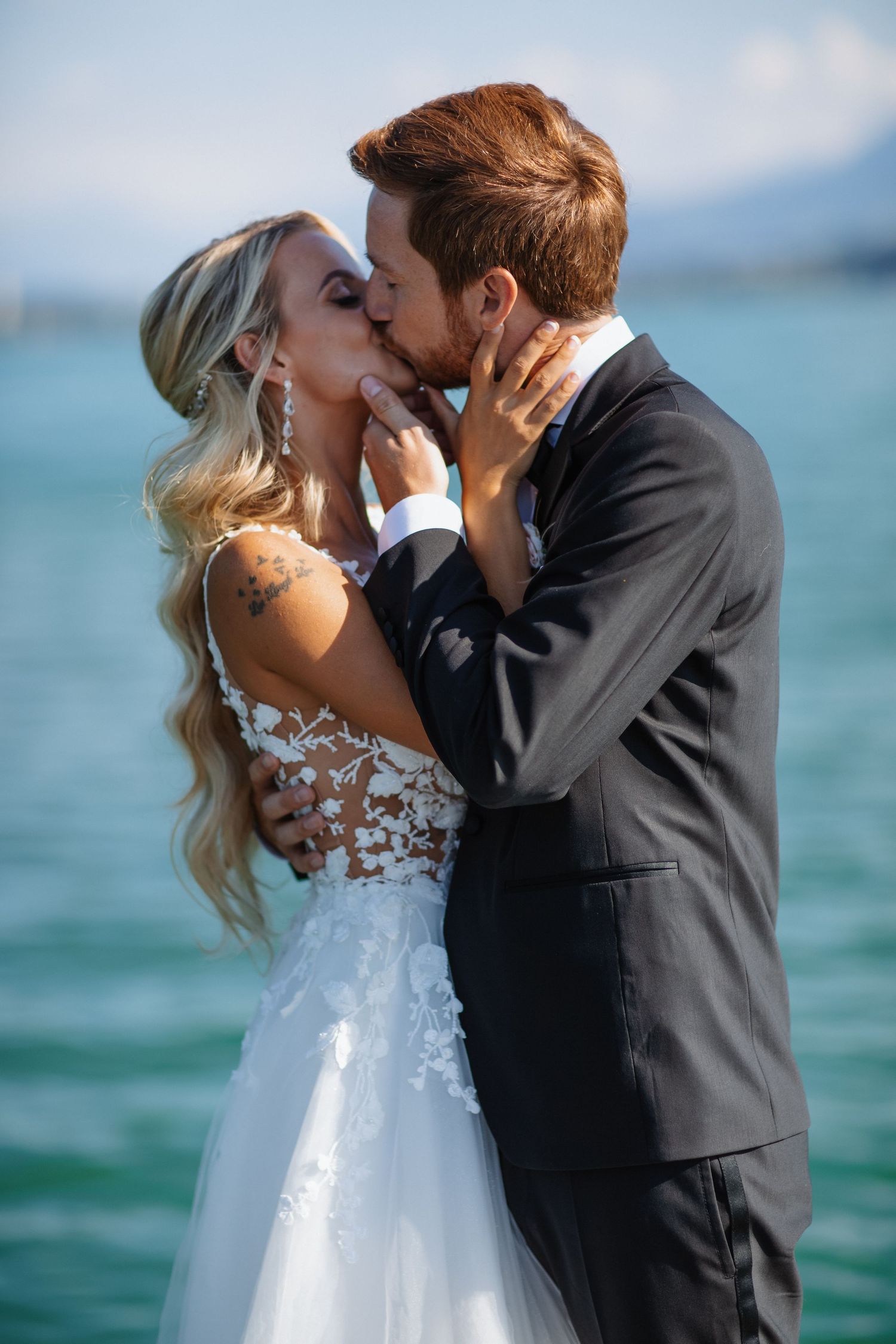 A bride and groom share a romantic kiss by the water, she in a lace gown, he in a dark suit.