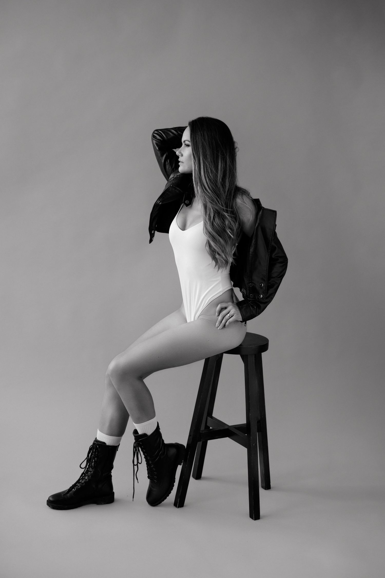 Black and white photo of a woman in a white bodysuit, leather jacket, and combat boots sitting on a stool.
