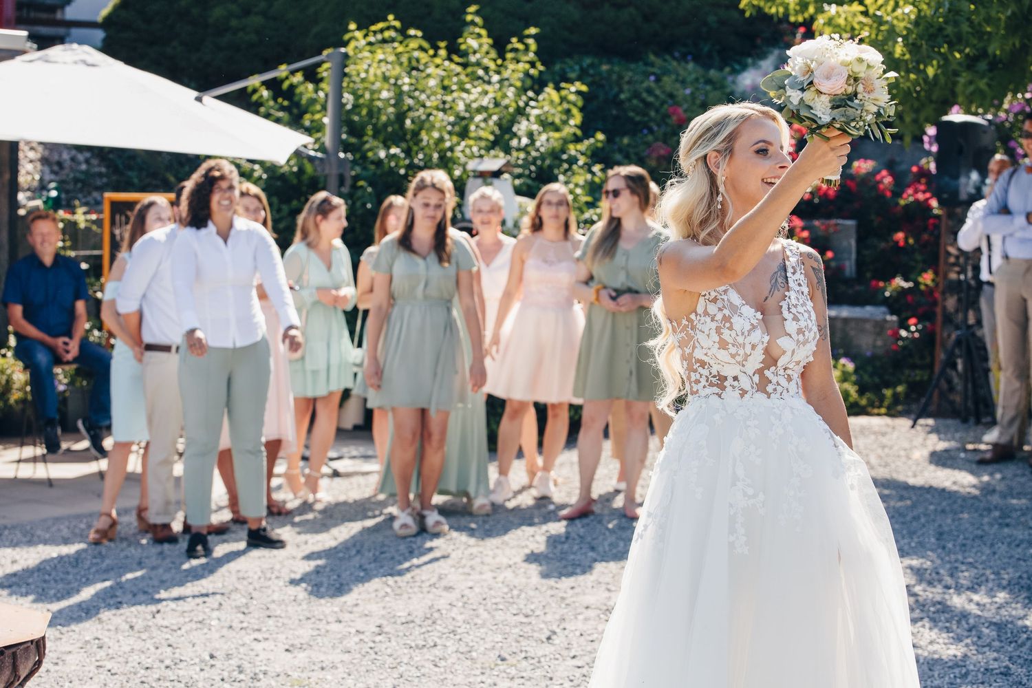 Bride in white lace gown tosses bouquet to group of guests in sage green dresses at outdoor wedding.