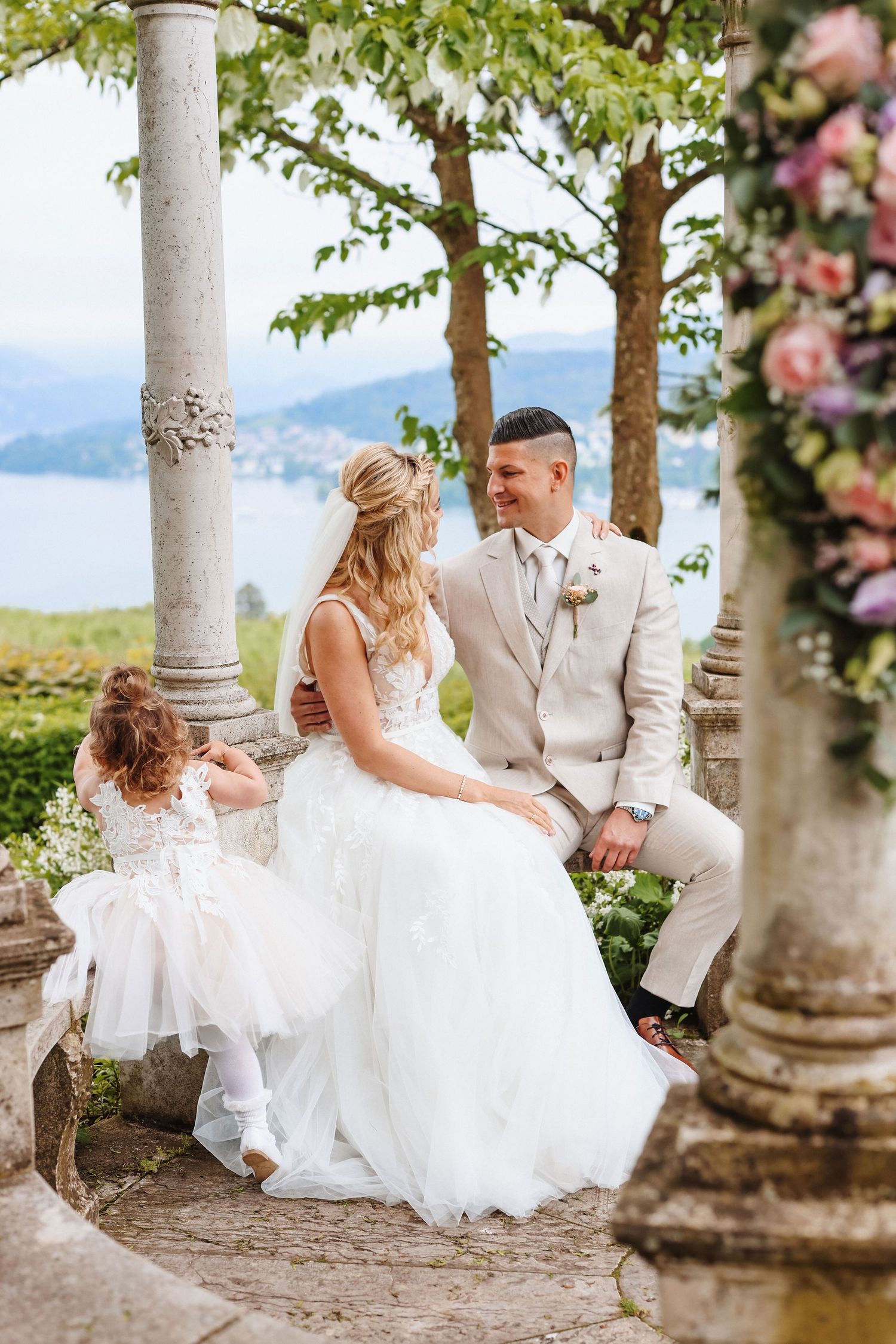 Bride and groom sit with flower girl under stone gazebo, scenic lake and mountains visible in background.