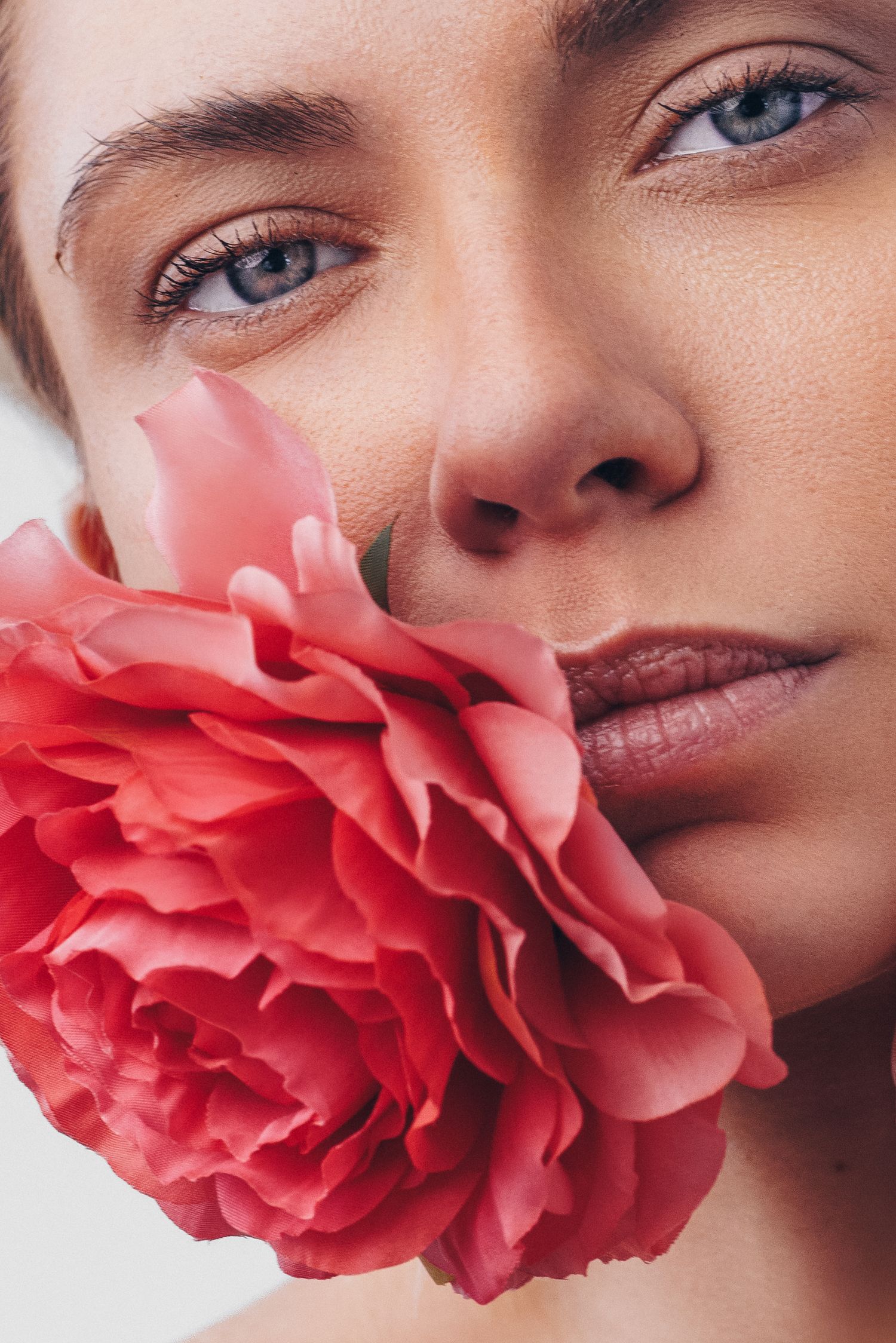 Close-up portrait of a woman with blue eyes holding a pink peony flower against her cheek.