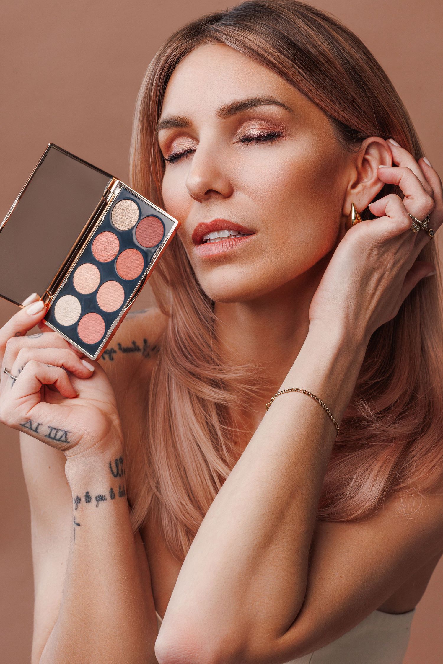 Woman with rose gold hair holds an eyeshadow palette, showcasing warm nude tones, with tattoos and gold jewelry visible.