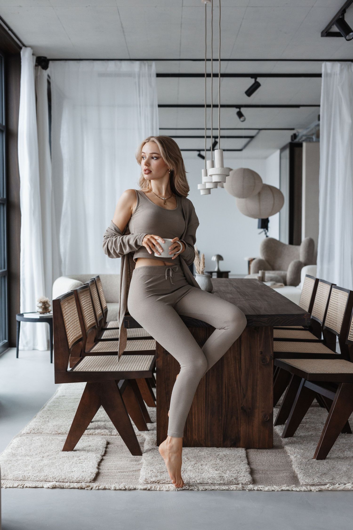 Woman in beige loungewear set sitting on wooden dining table holding coffee cup in modern interior.