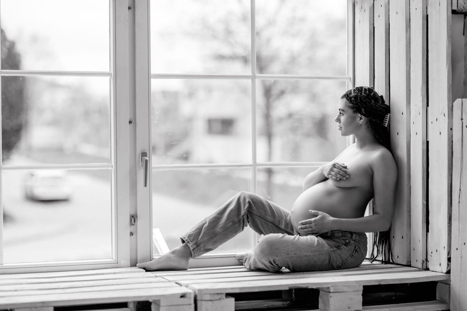 Black and white photo of a pregnant woman sitting by a window, cradling her bare belly in soft natural light.