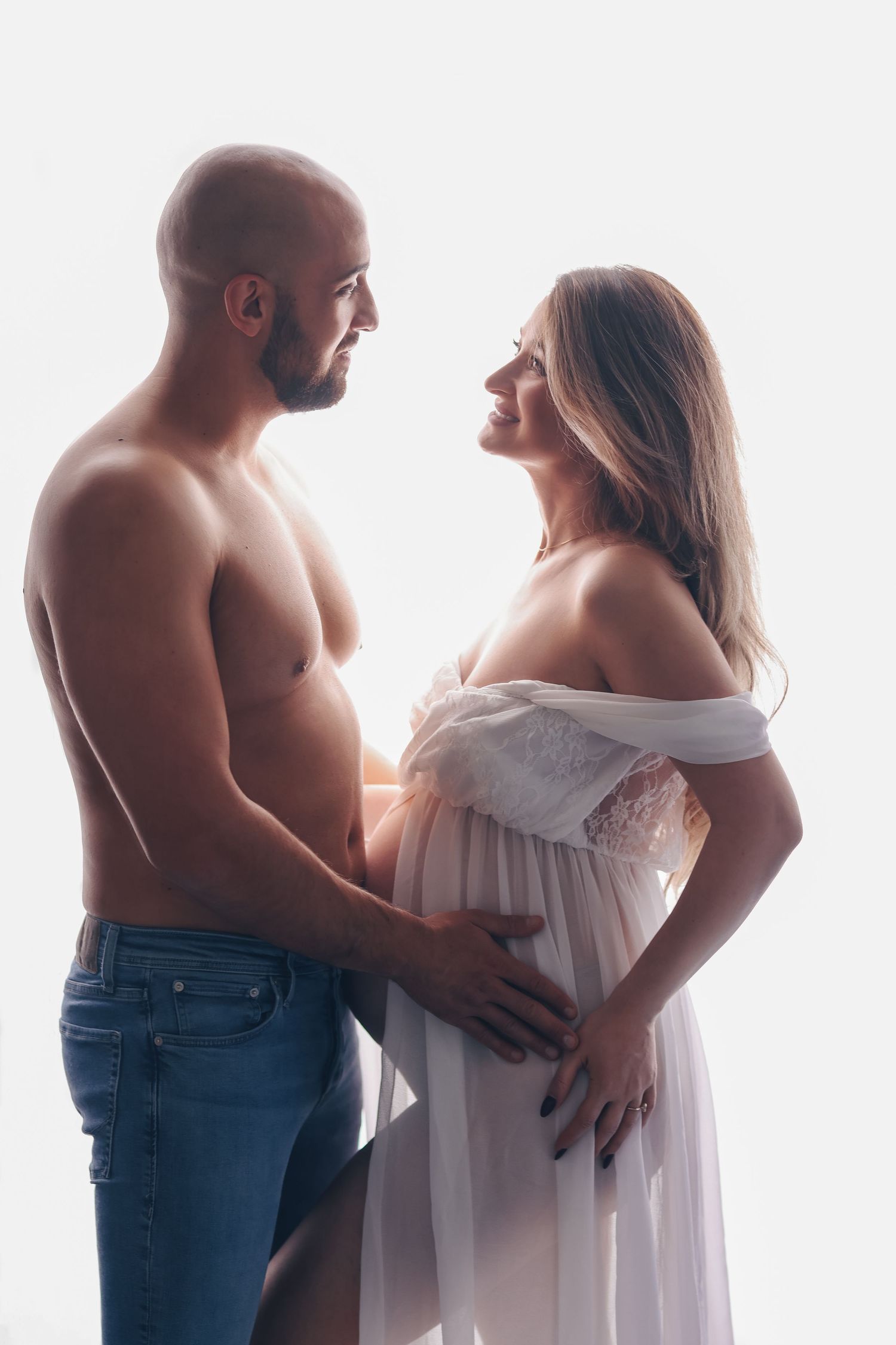 Shirtless bald man and smiling pregnant woman in white dress embracing each other in a bright studio setting.
