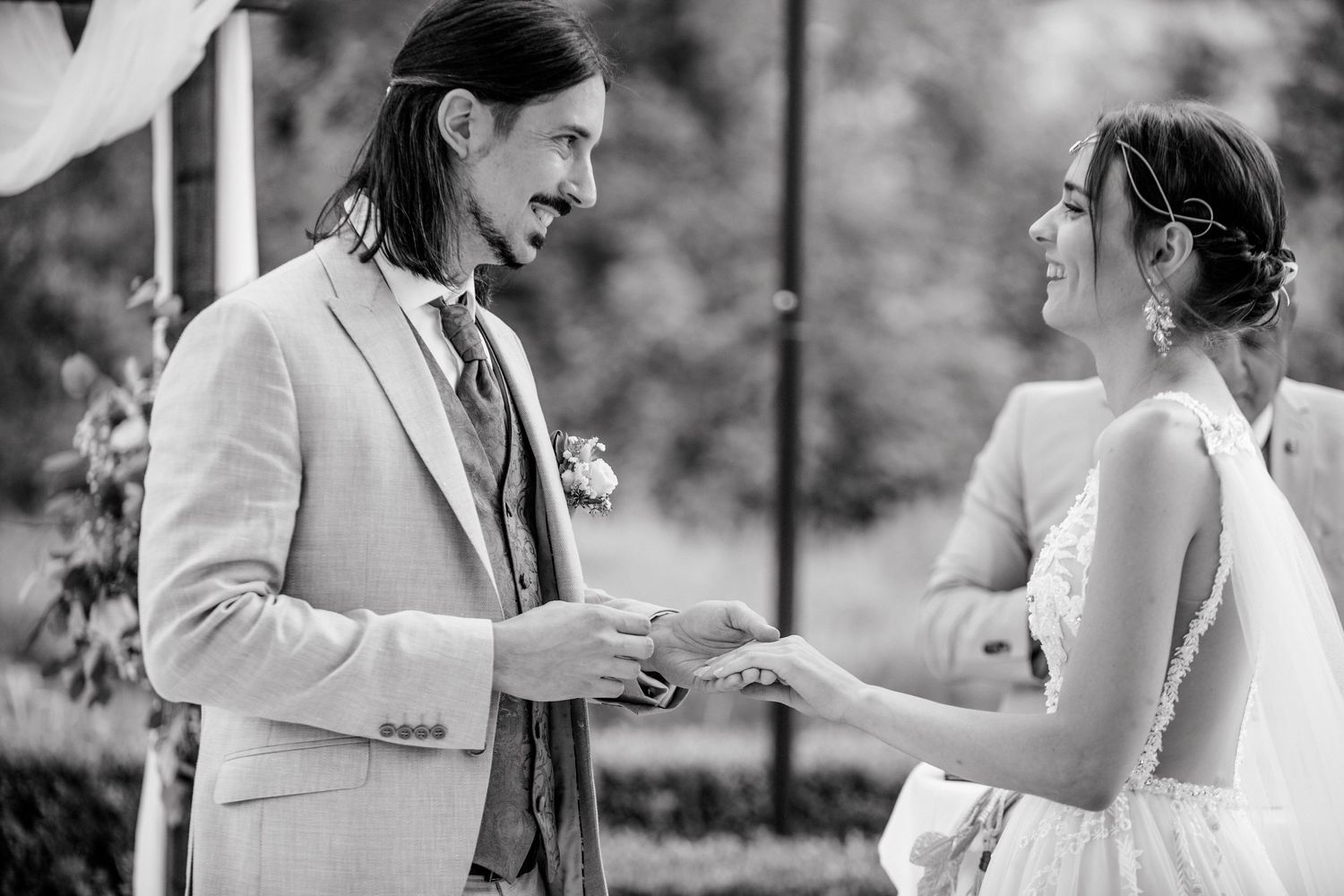 Black and white wedding photo of groom in suit exchanging rings with smiling bride outdoors.